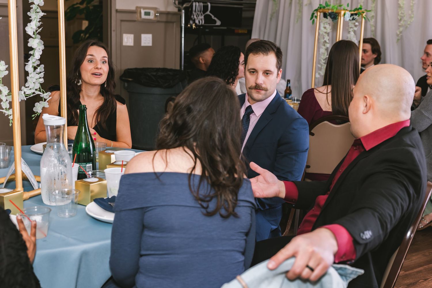 Guests in formal attire conversing at a round wedding reception table with champagne and dinnerware.
