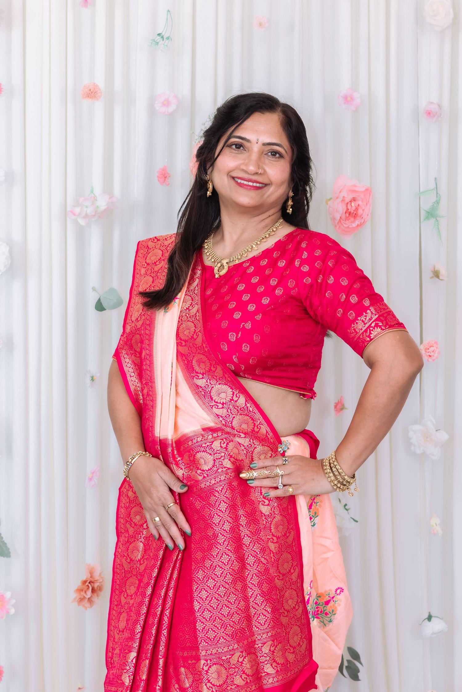 Smiling Indian woman in pink Banarasi saree with gold jewelry poses at a floral decorated white wedding backdrop.