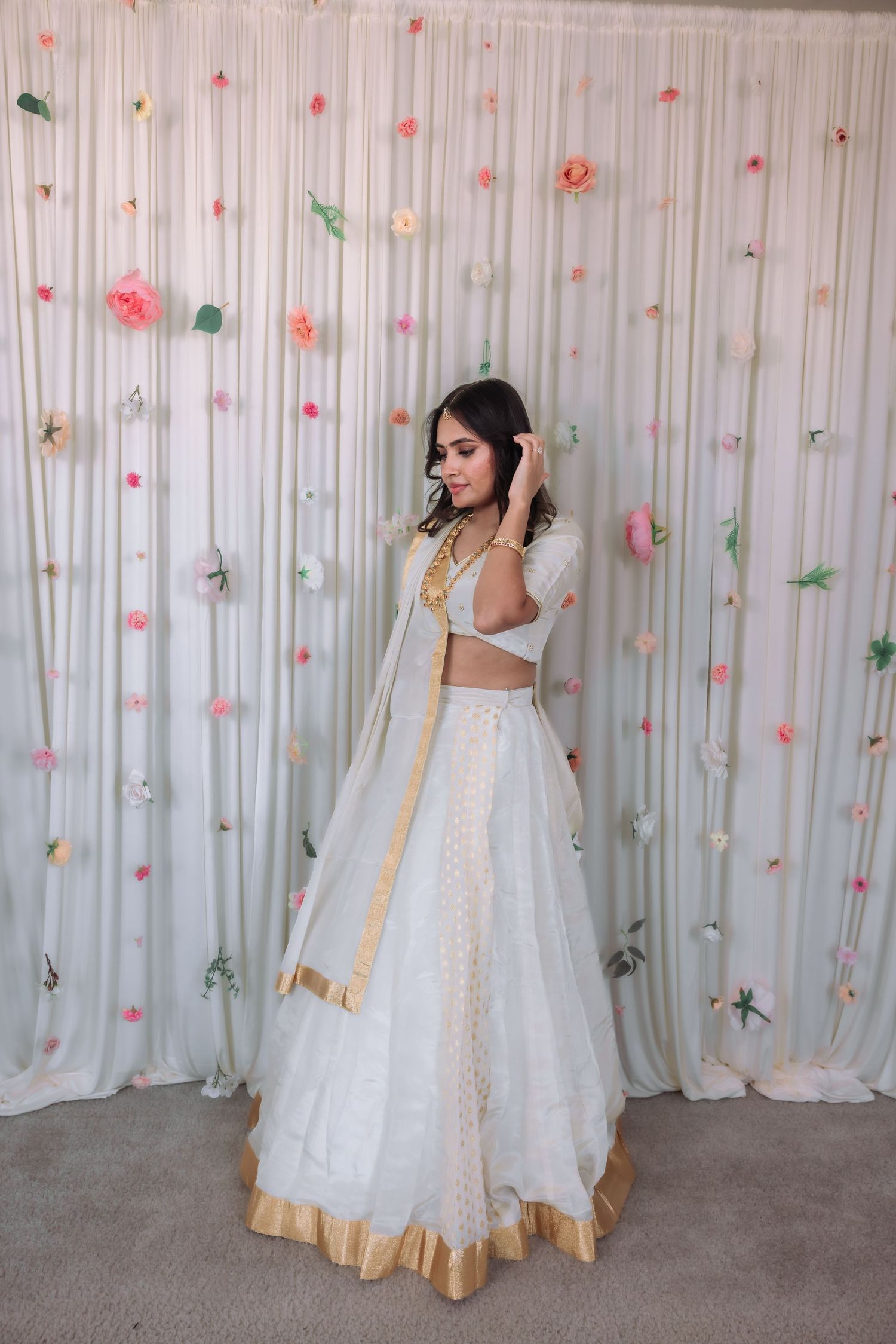 Bride in white and gold lehenga poses gracefully before floral curtain backdrop at Indian wedding.