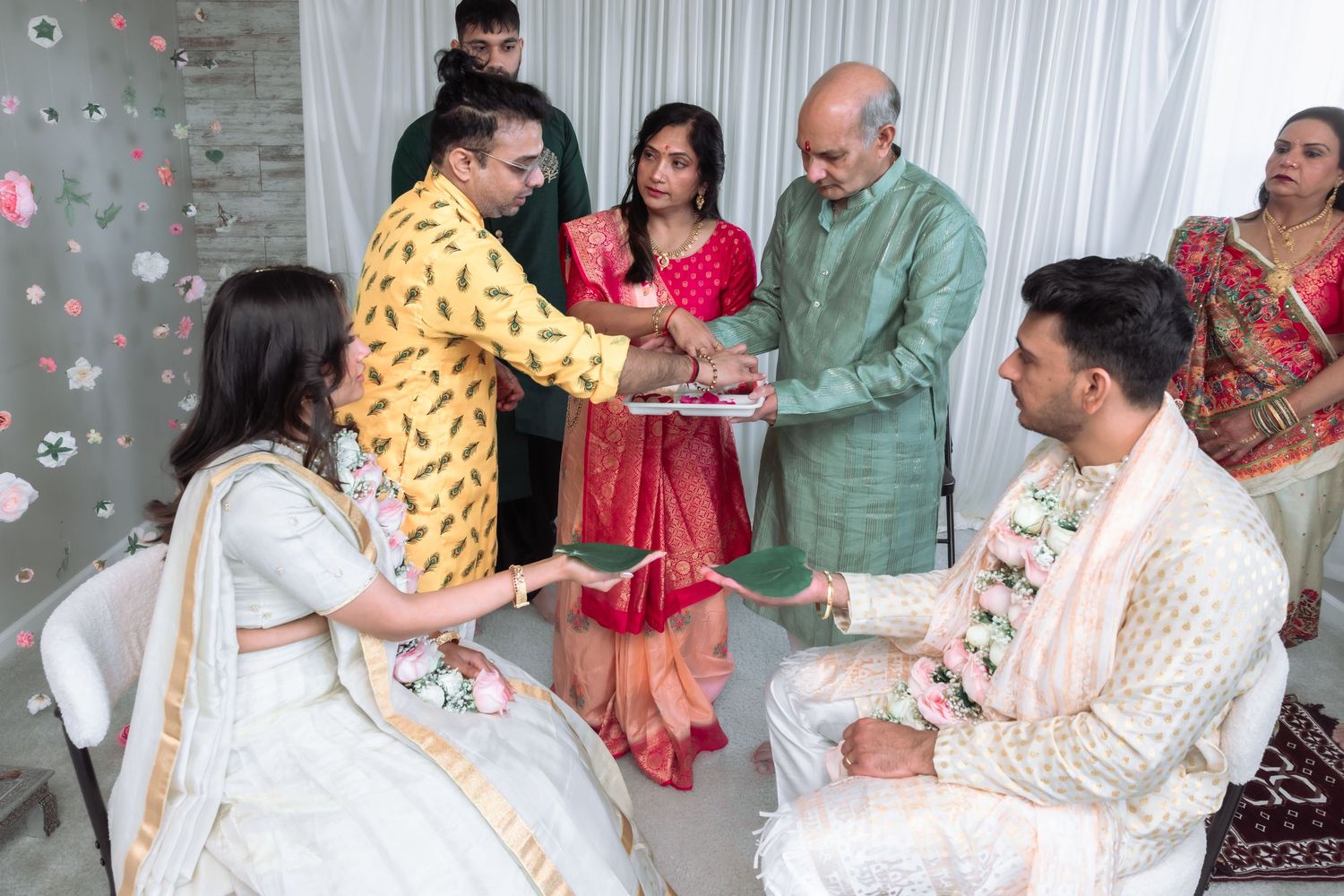 Family members exchange gifts during a traditional Indian wedding ceremony with guests in colorful ethnic attire.