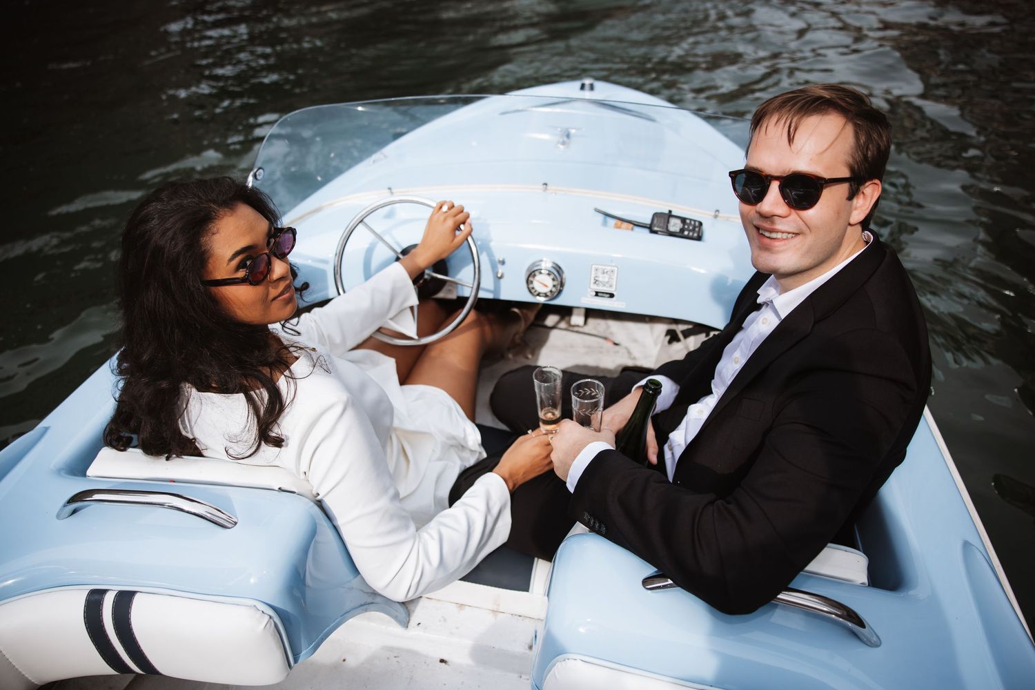 Couple smiling and toasting champagne on a light blue vintage boat on a scenic city waterway.