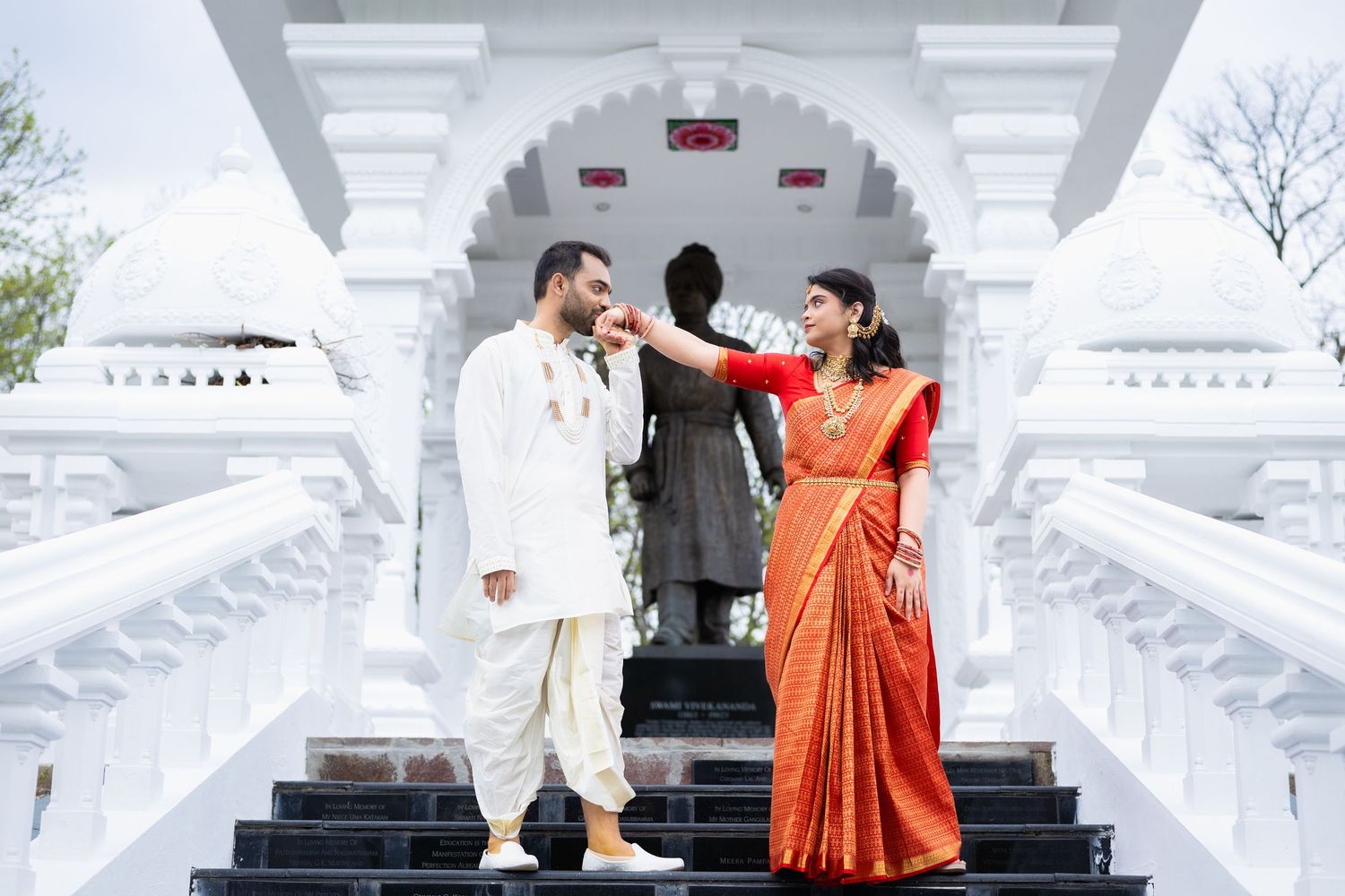 Indian bride in red saree and groom in white kurta posing at ornate white Hindu temple steps.