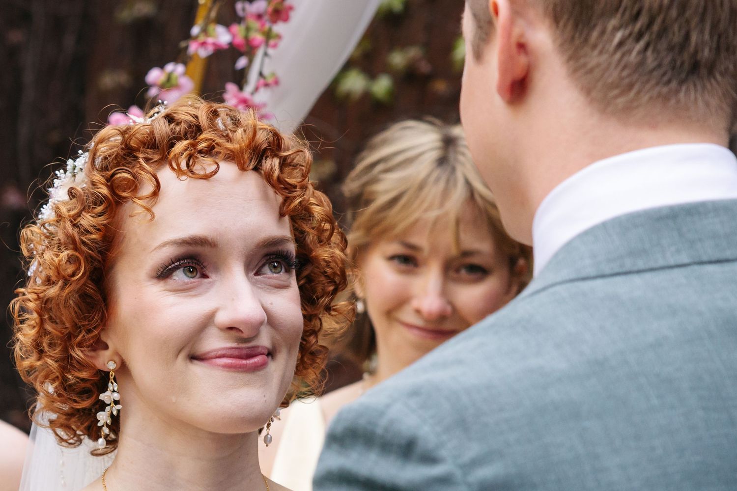 Bride with curly red hair smiles at groom during outdoor wedding ceremony with officiant in background.