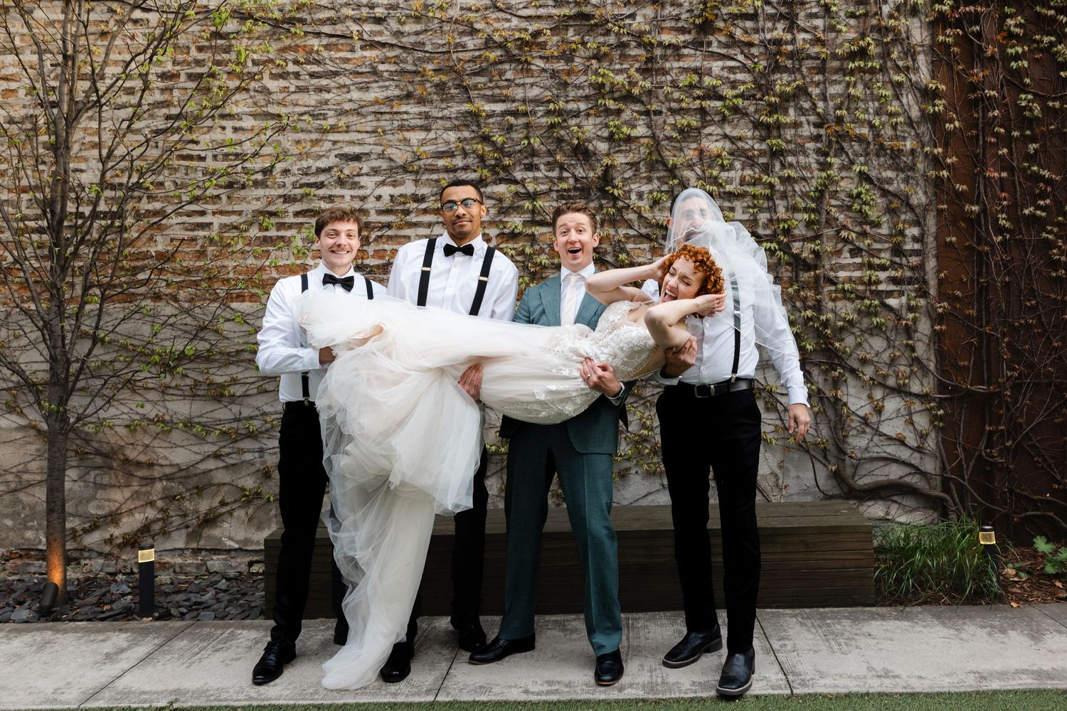 Groom and groomsmen in suspenders playfully lift the bride in a white gown outdoors against a brick wall.