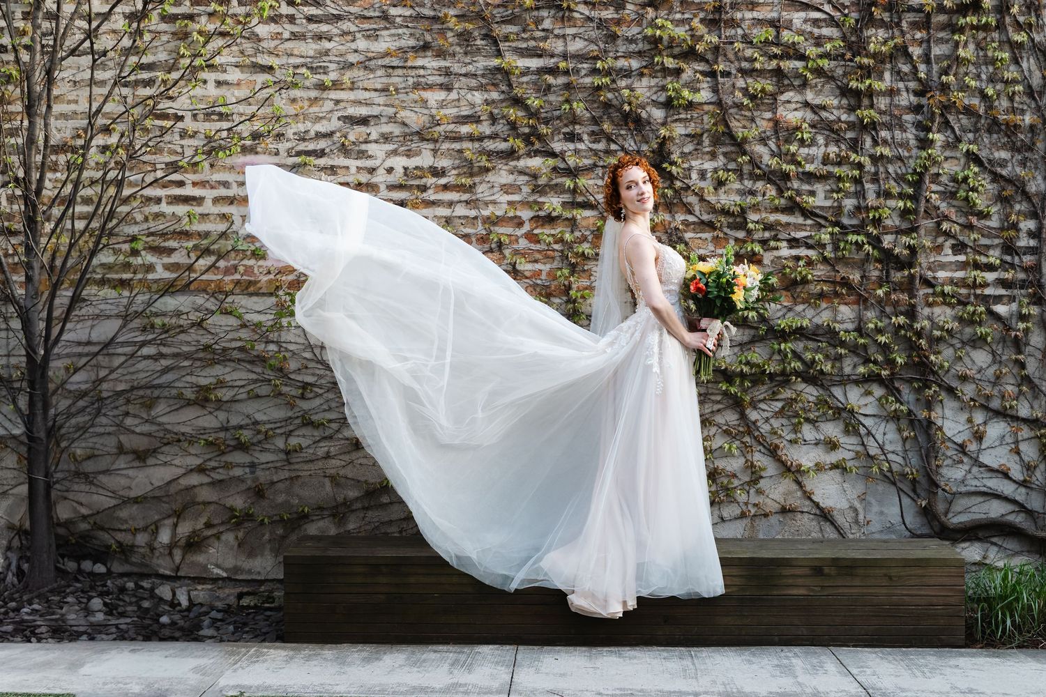 Redhead bride in flowing white gown tosses skirt while holding colorful bouquet against ivy-covered brick wall.