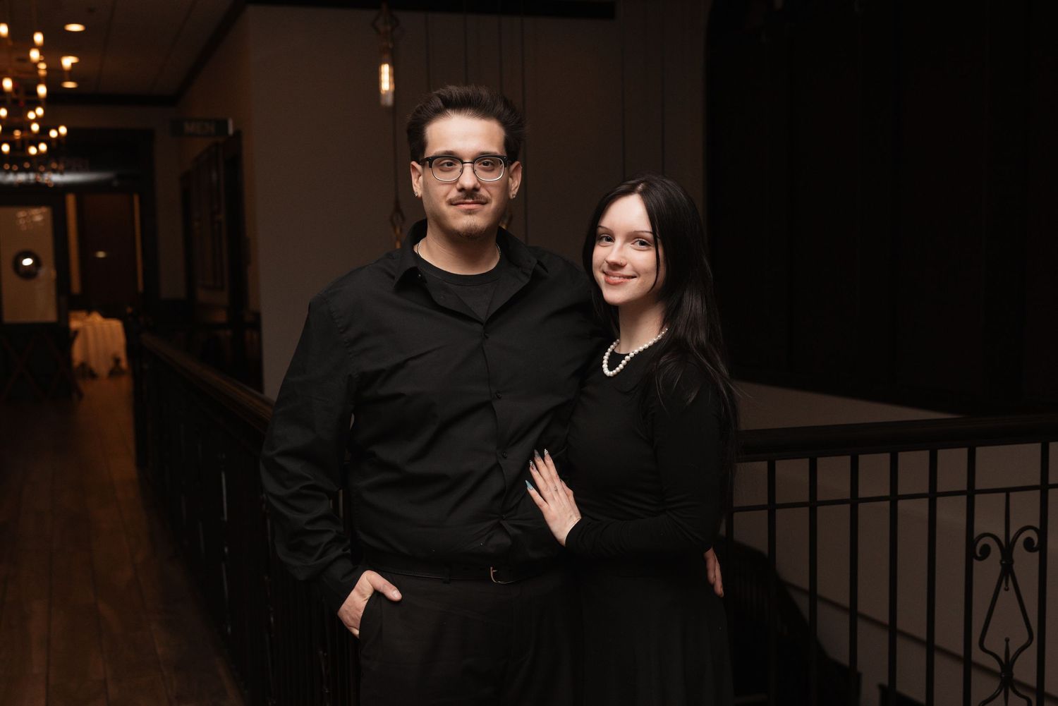 Couple in matching black outfits pose together in an elegant dimly lit venue.
