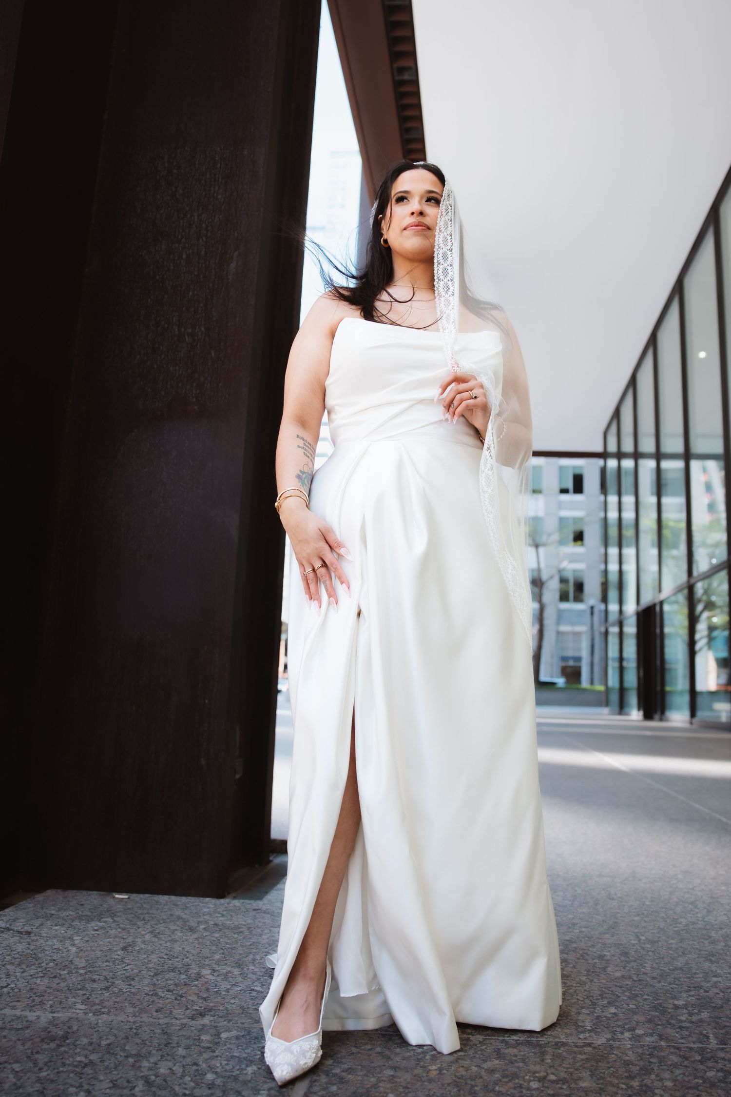 Bride in white strapless gown with veil posing near dark column outdoors.