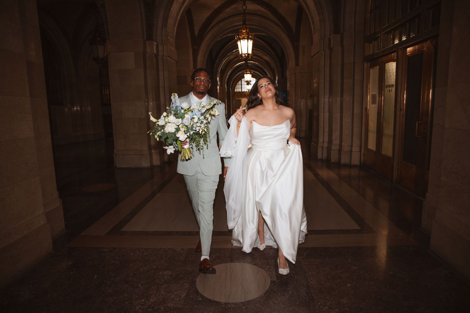 Bride and groom walking through elegant arched hallway, bride in white gown holding dress, groom in light suit with bouquet.