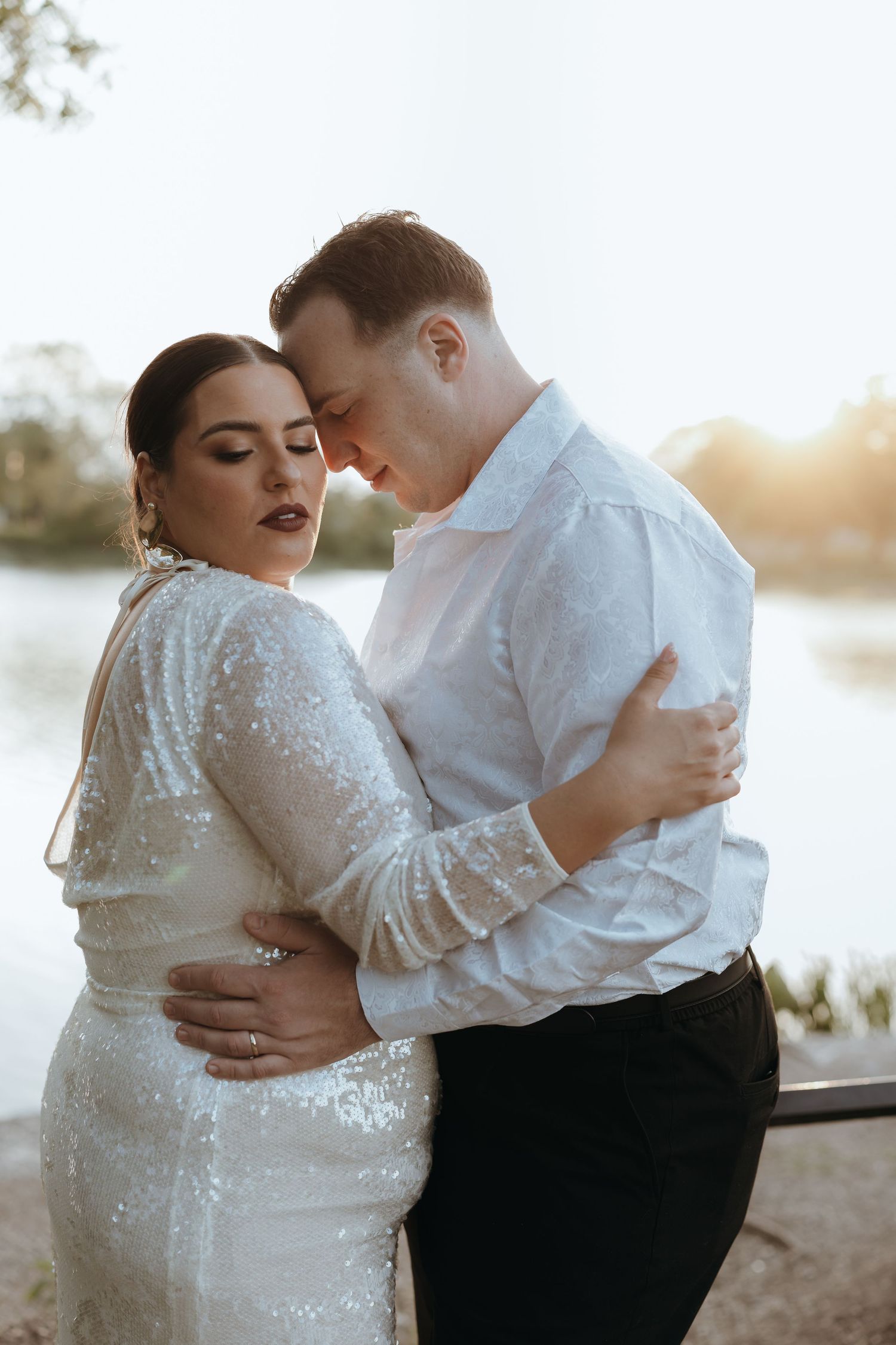 Bride and groom embrace tenderly by waterside at golden hour in sequined wedding dress.