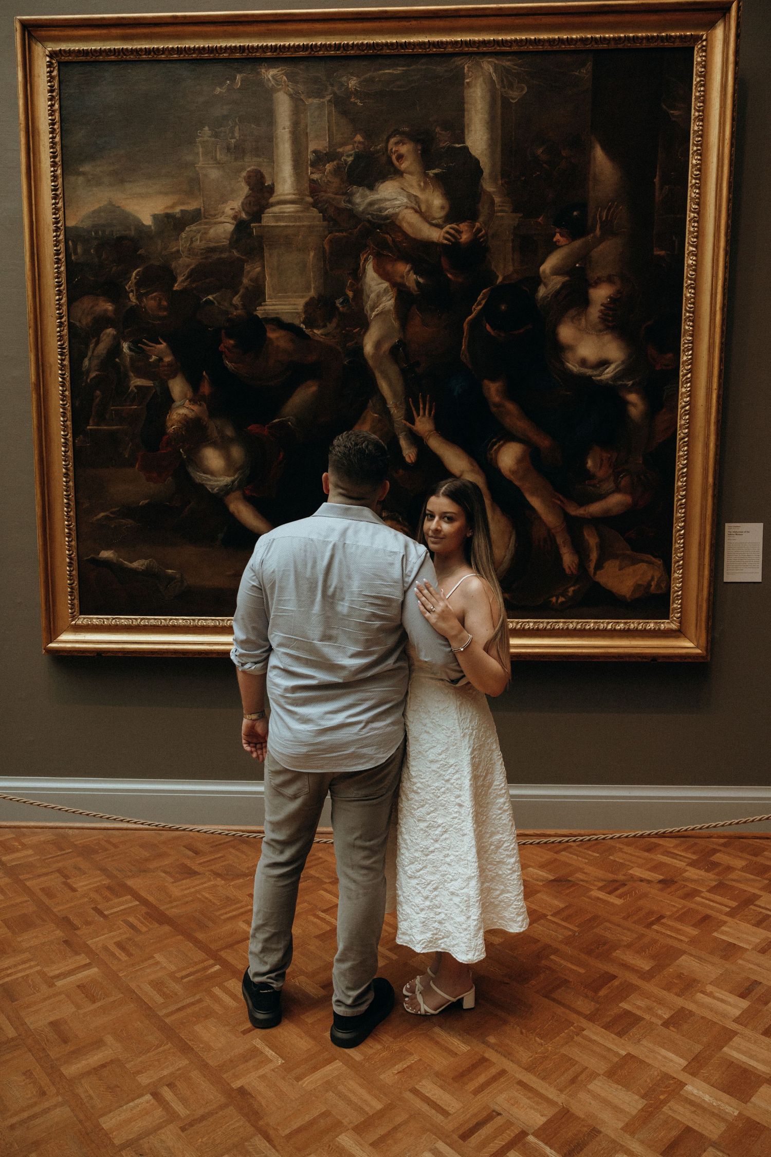 Couple stands hand-in-hand admiring a large classical painting at an art museum engagement session.