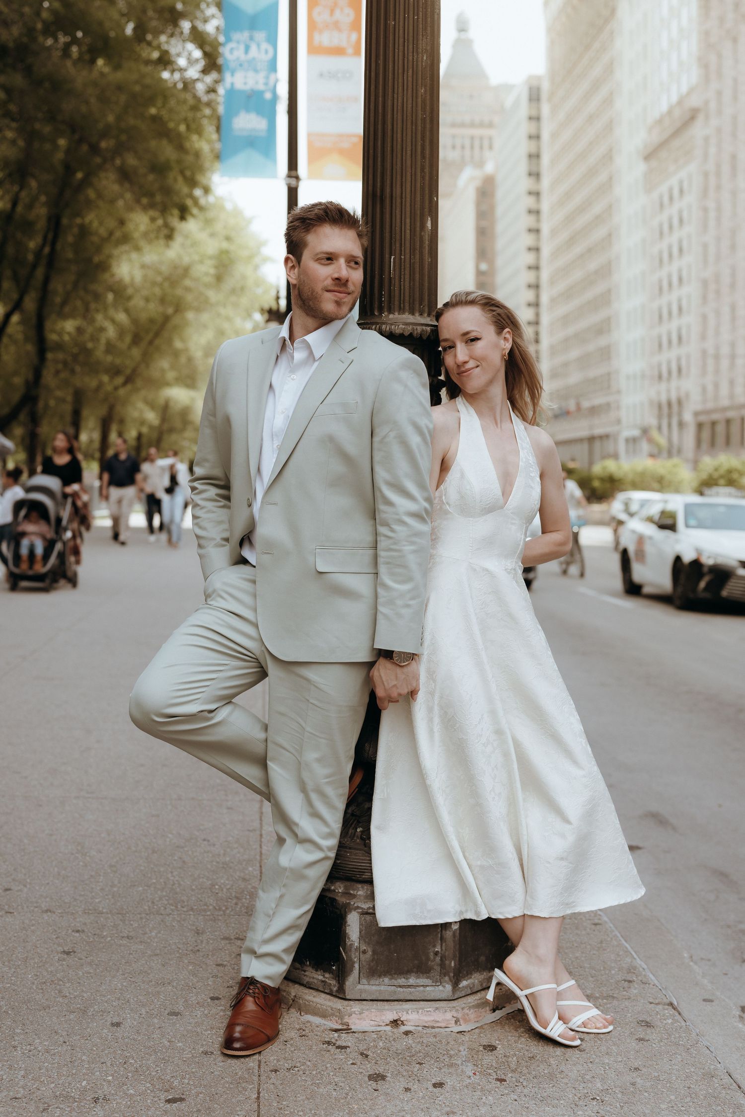 Bride and groom pose against a lamppost on a city street, she in white dress, he in beige suit.