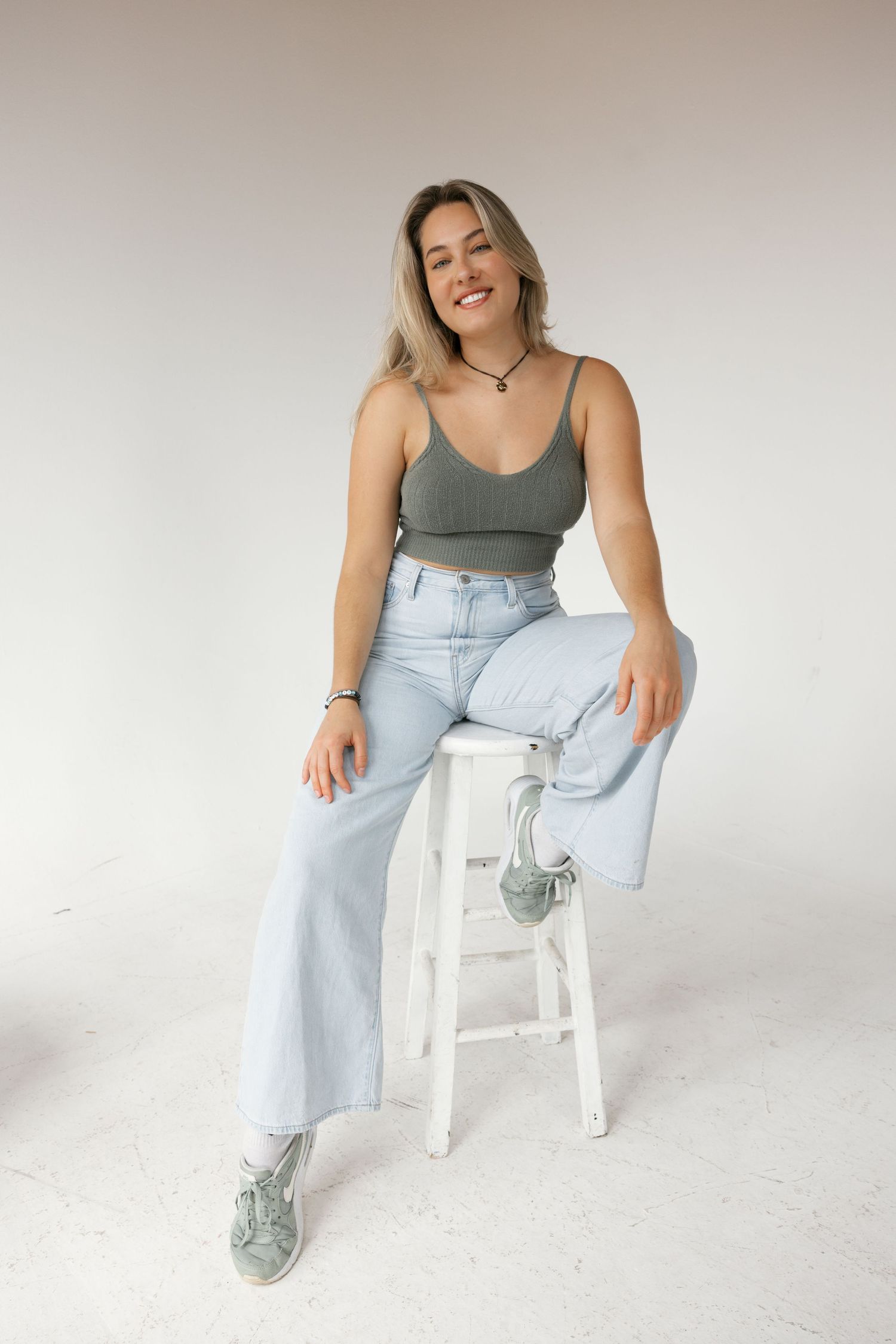 Smiling blonde woman in gray crop top and wide-leg jeans sitting on white stool in studio.