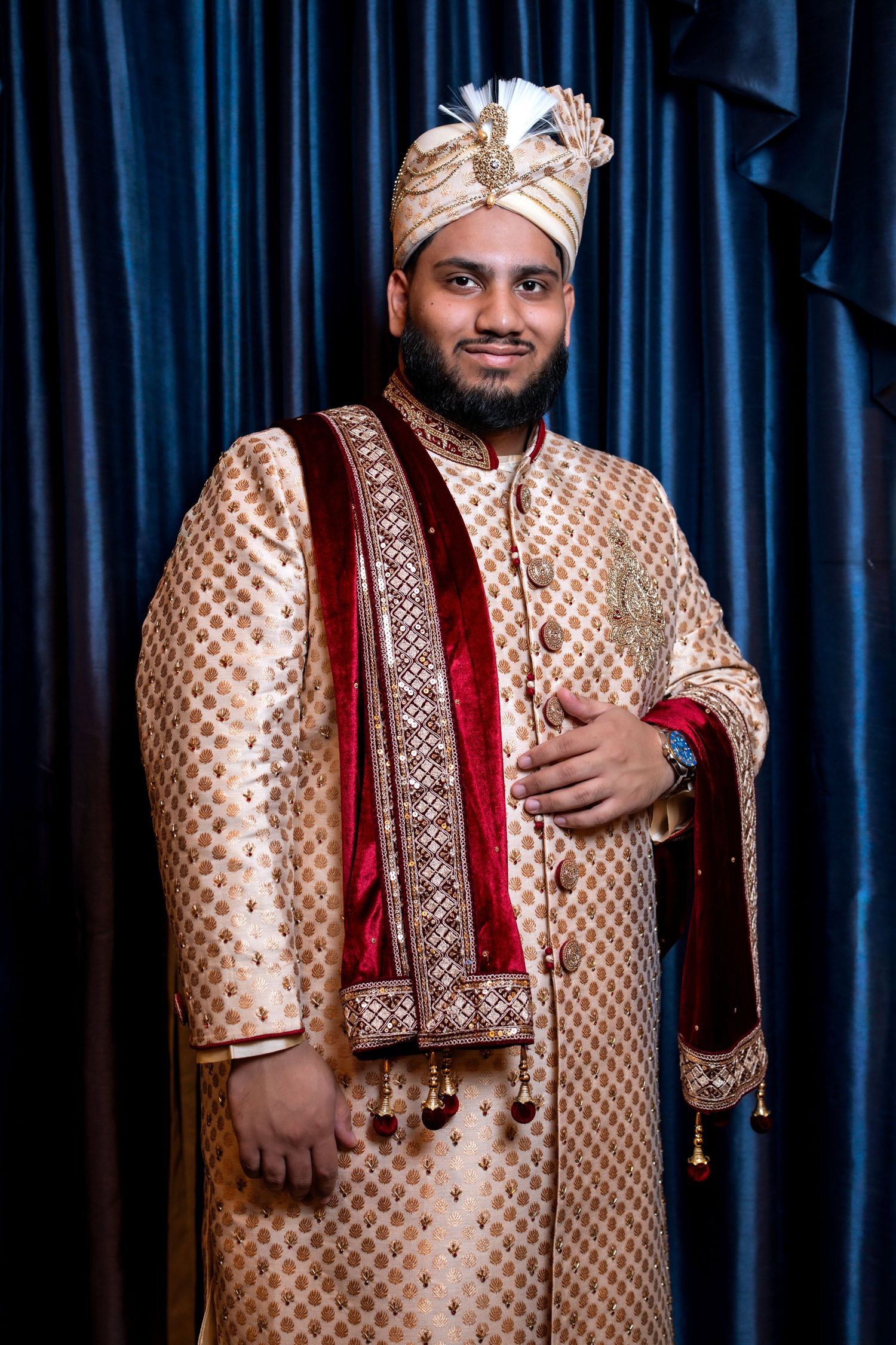 Indian groom in ornate gold sherwani with red velvet stole and turban smiling before blue curtains.