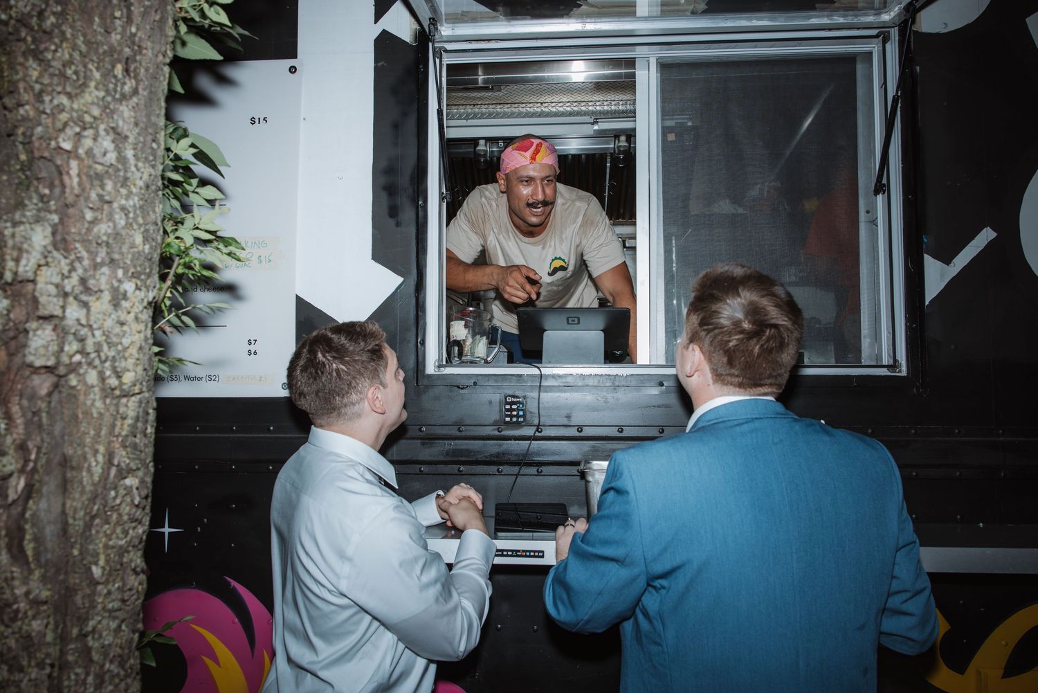 A food truck worker hands food to two men in suits through a service window at night.