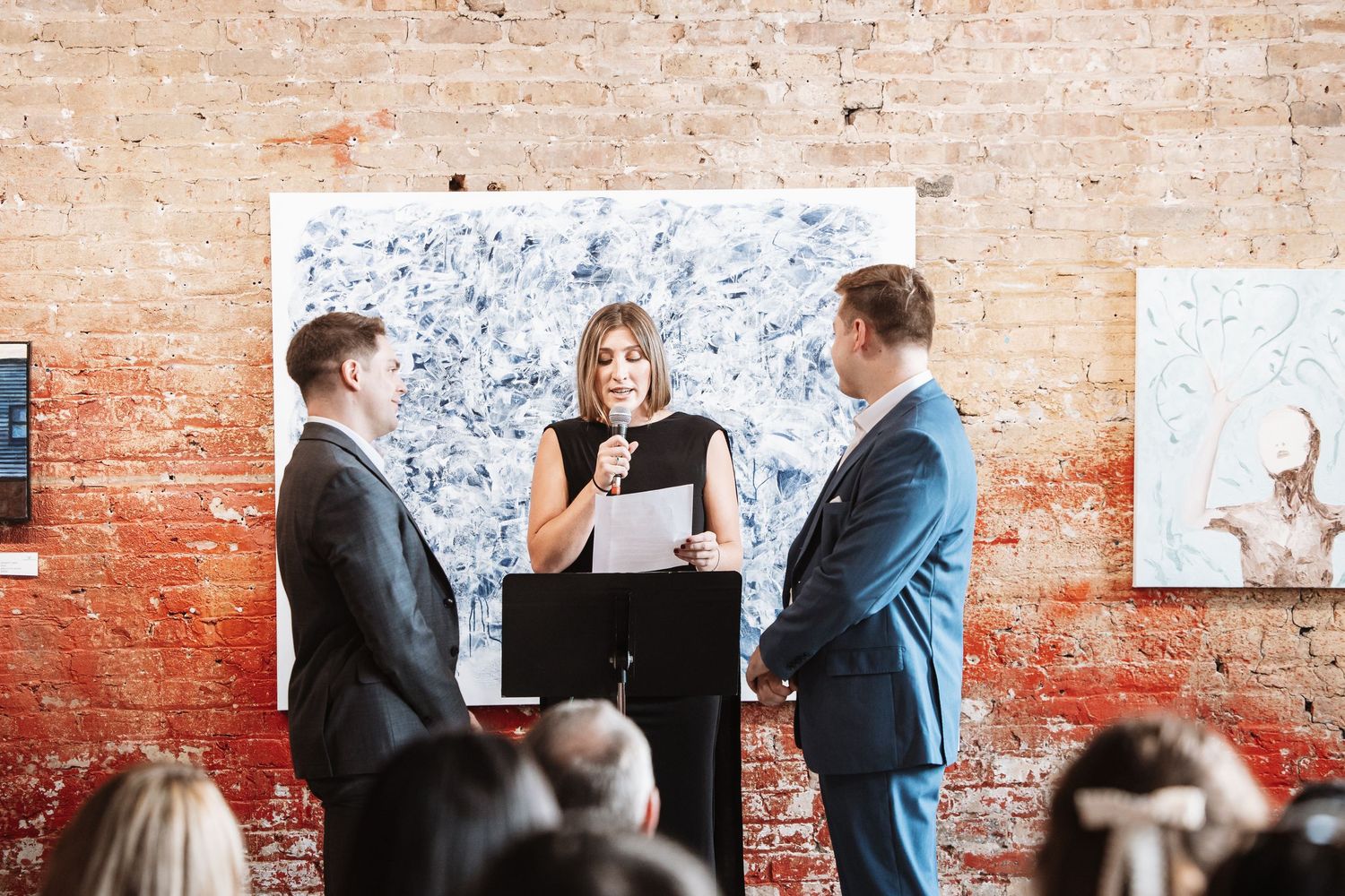 Wedding officiant with microphone reading to couple standing in brick wall venue.