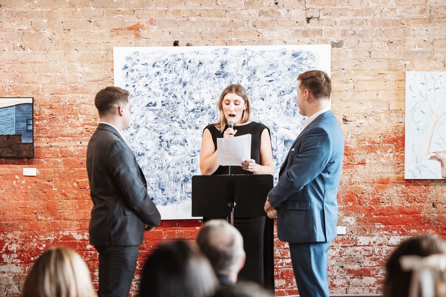Woman reads from paper at podium between two men in suits at an art gallery ceremony.