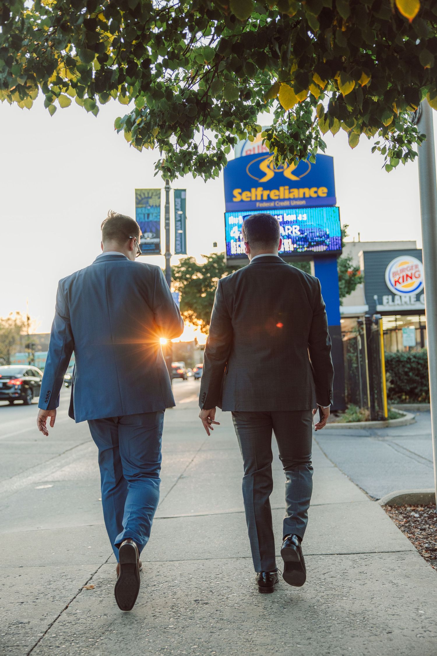 Two men in suits walking on sidewalk past Selfreliance credit union sign at sunset.