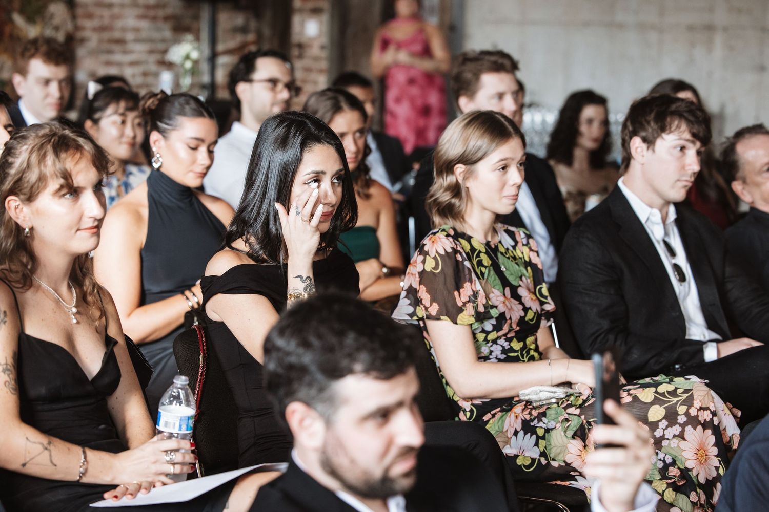 Diverse group of guests seated at an indoor wedding ceremony, with one woman wiping a tear.