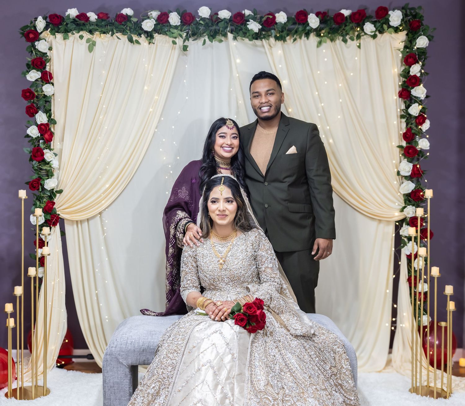 Bride in gold lehenga holds red roses posing with two guests at decorated floral wedding backdrop.