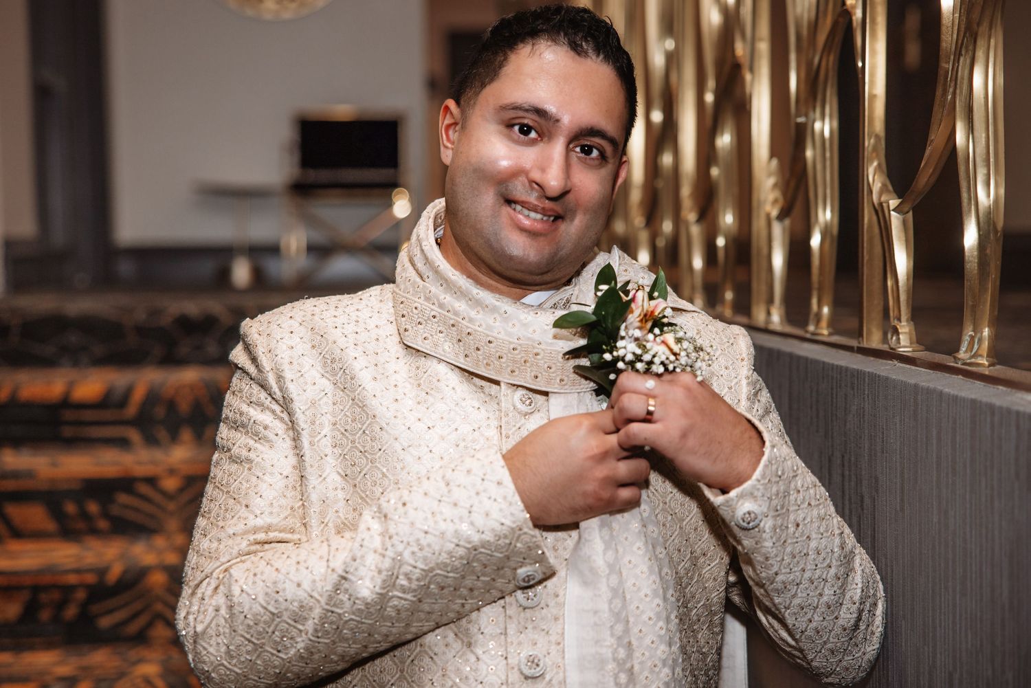 Smiling groom in cream sherwani adjusts floral boutonniere near golden decorative railing.