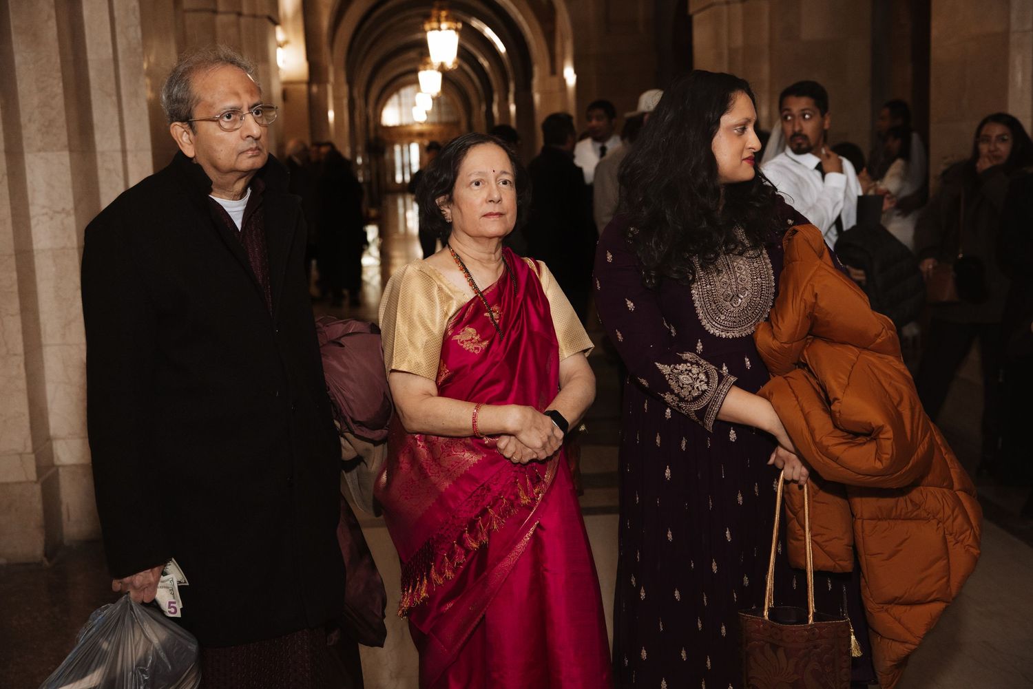 Three elegantly dressed wedding guests walk through ornate cathedral corridor wearing traditional Indian and formal attire.