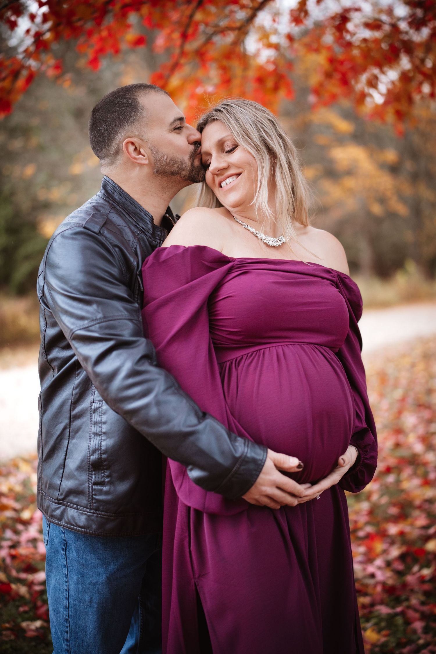 Man kissing pregnant woman's cheek as she smiles, both holding baby bump in autumn park.