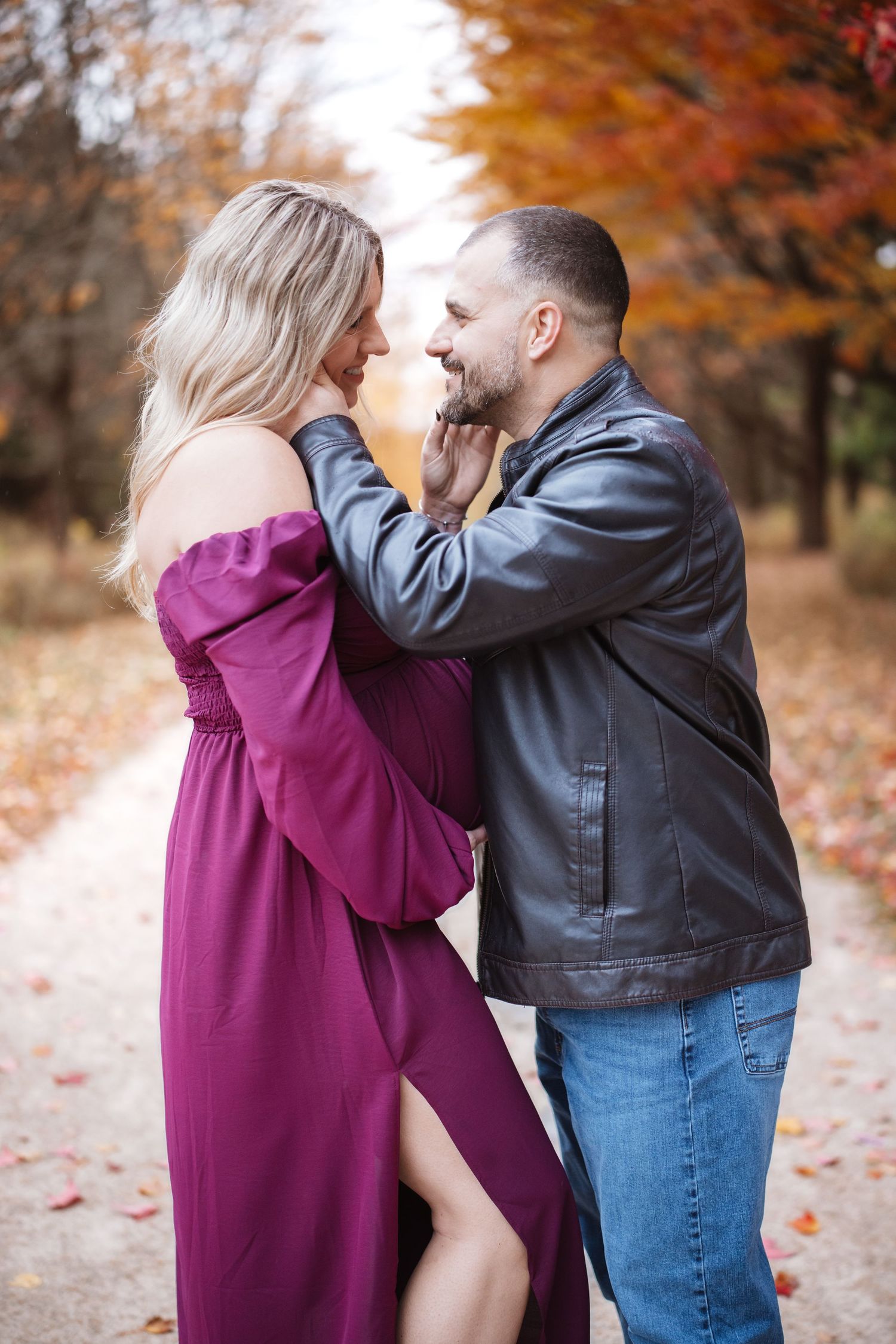 Couple sharing romantic moment on autumn path, man in leather jacket tenderly holds womans chin as she smiles in purple dress.
