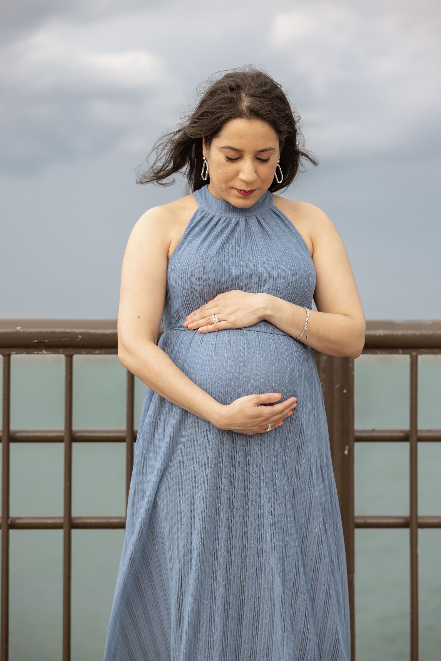 Pregnant woman in blue halter dress gently holding her baby bump on a waterfront railing with overcast skies.