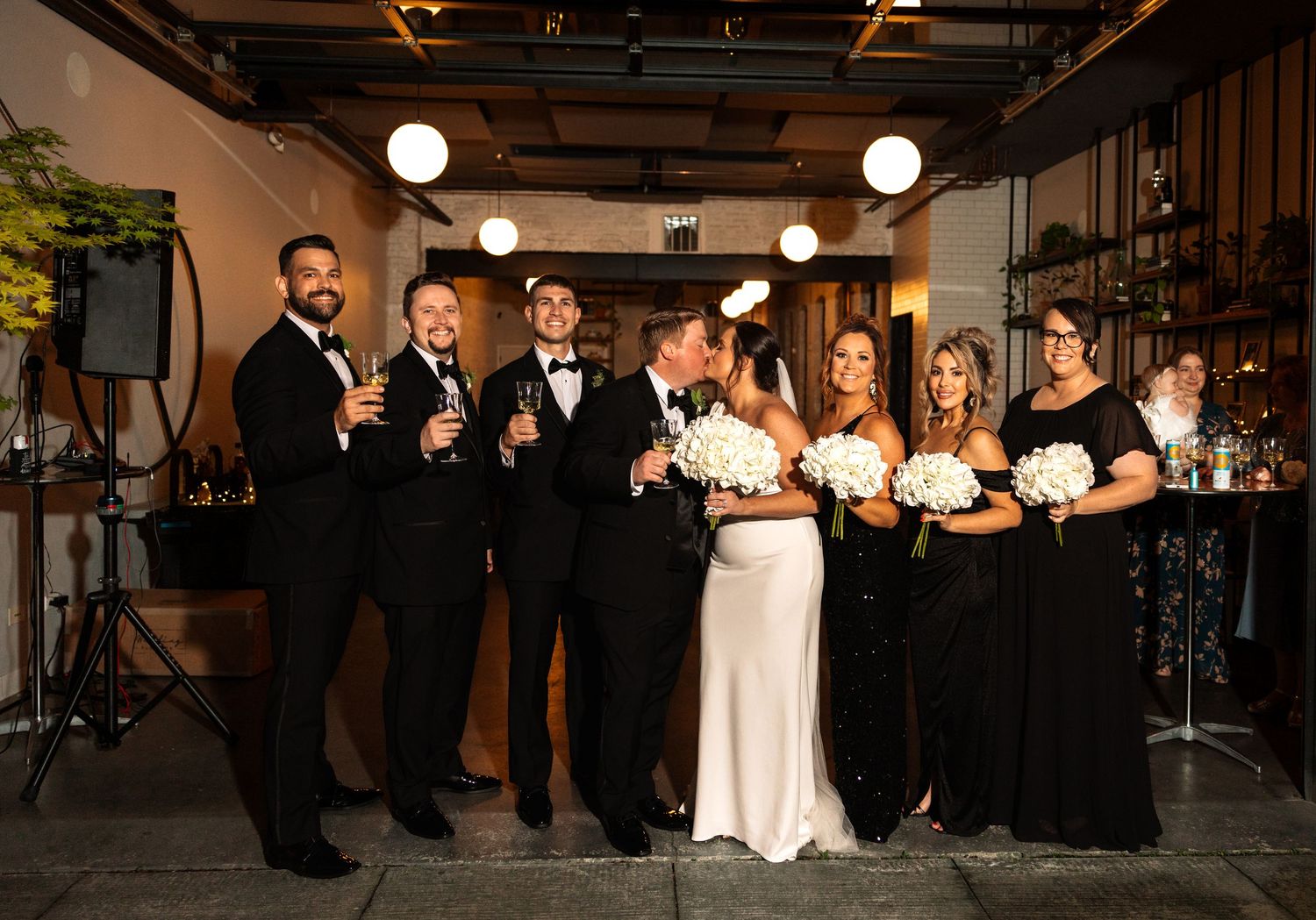 Wedding party celebrating with champagne in industrial venue, bride in white and party in black attire.