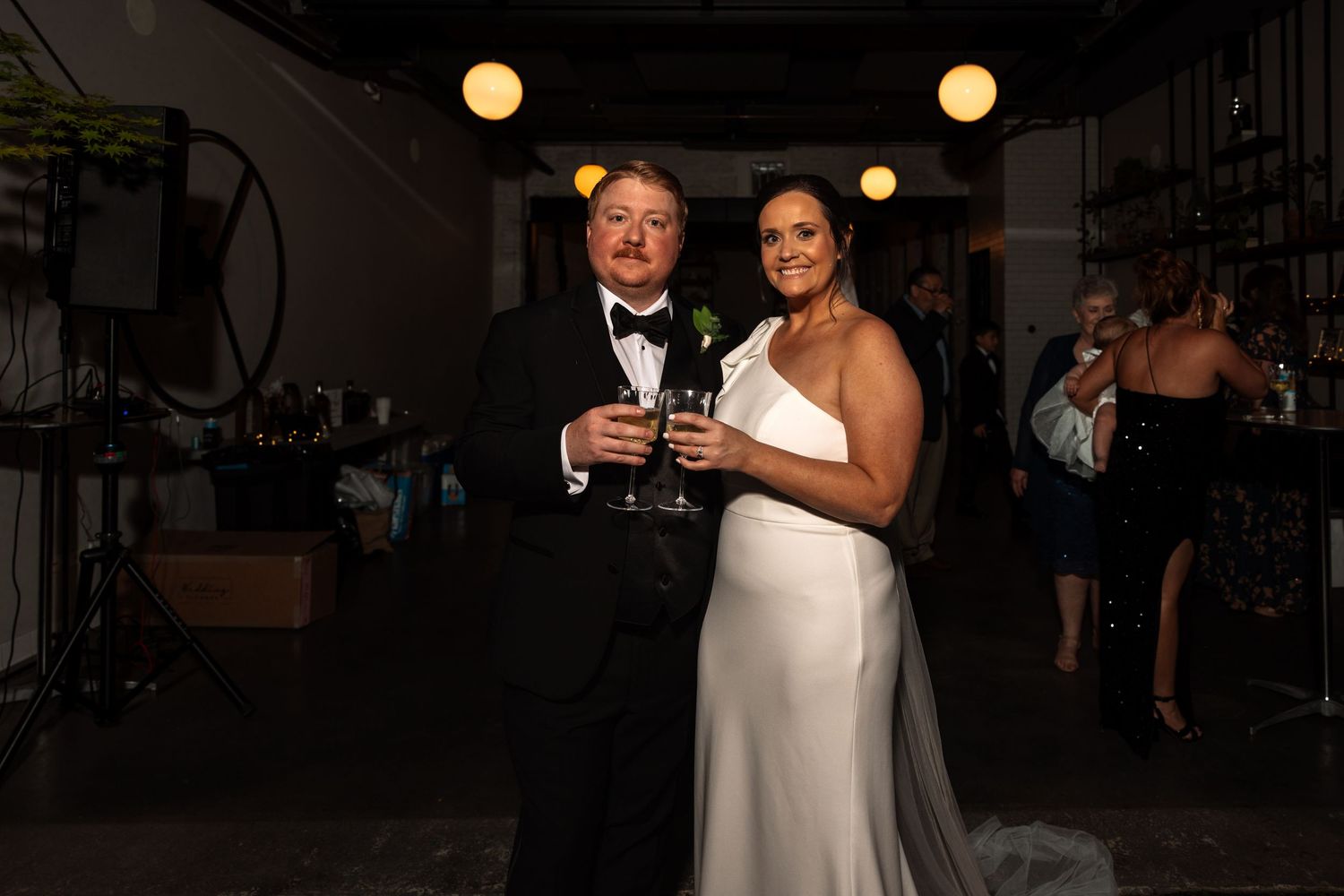 Bride and groom pose together holding champagne glasses at dimly lit wedding reception venue.