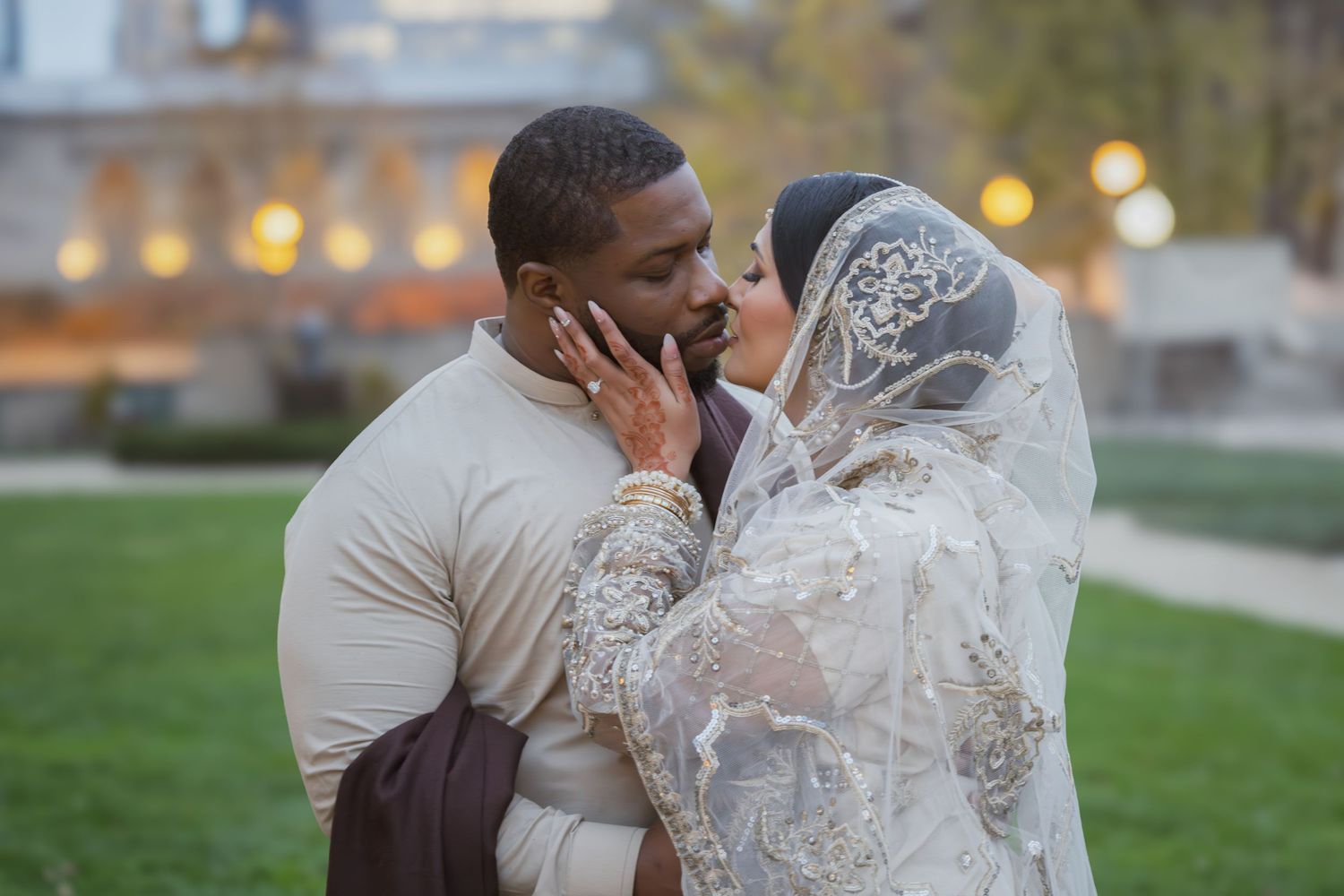 Interracial Muslim newlyweds sharing intimate moment outdoors, bride in ivory embroidered bridal attire with veil.