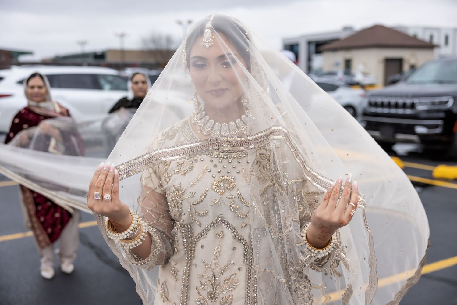 South Asian bride in ornate white lehenga holding flowing veil at outdoor wedding.
