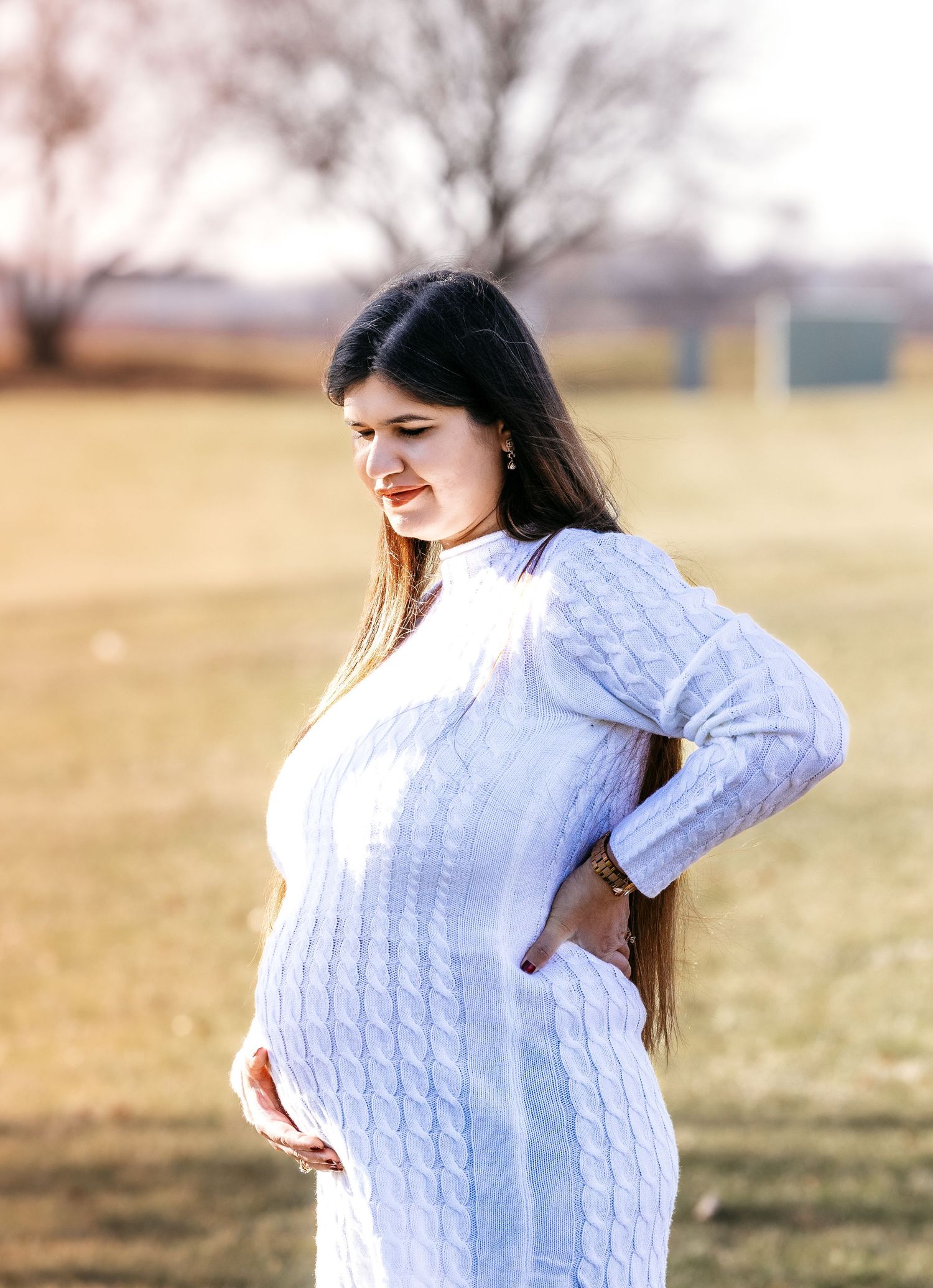 Pregnant woman in white cable-knit dress holding baby bump and back during outdoor maternity shoot.