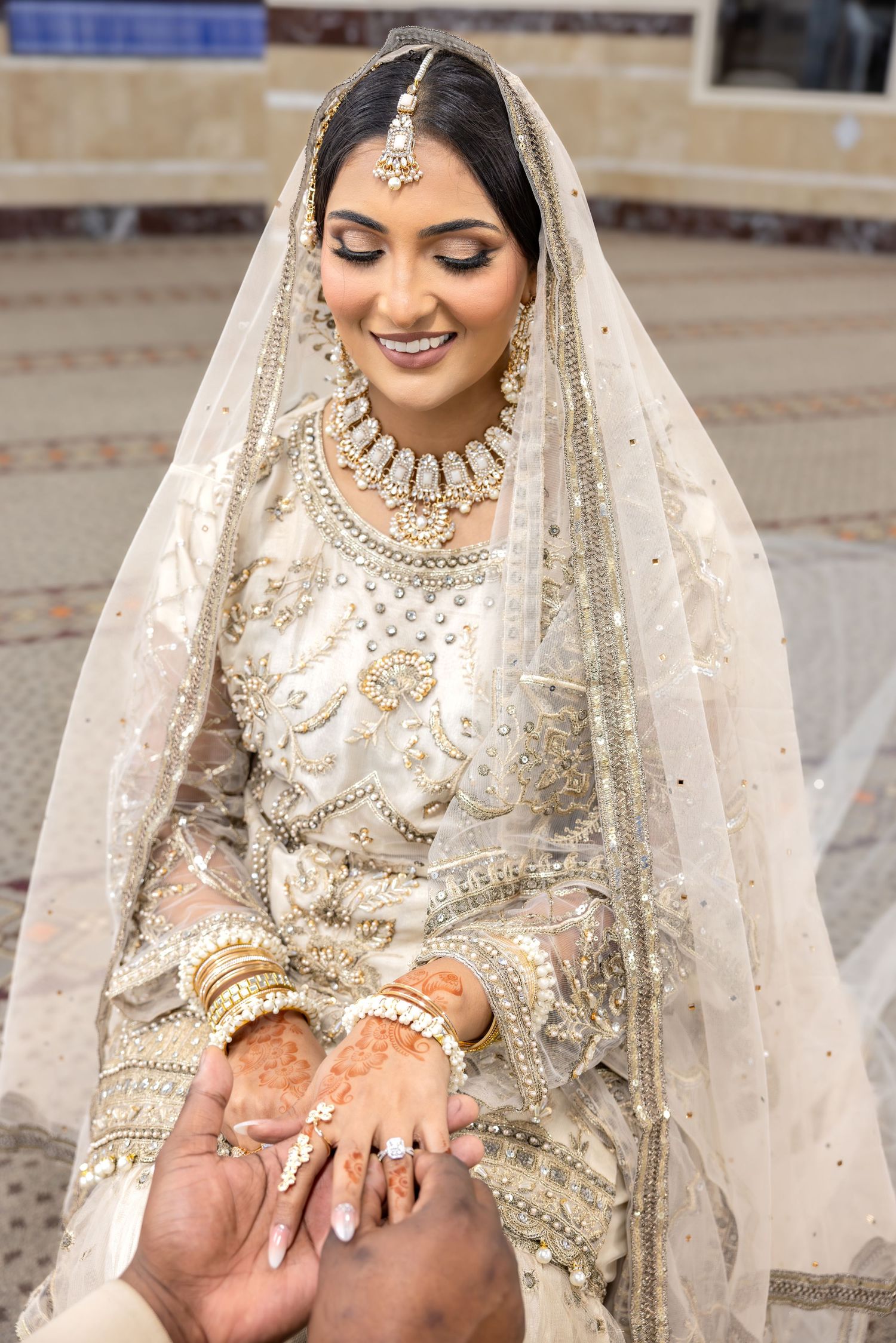 Beautiful South Asian bride in white gold embroidered bridal lehenga smiling while holding groom's hands.