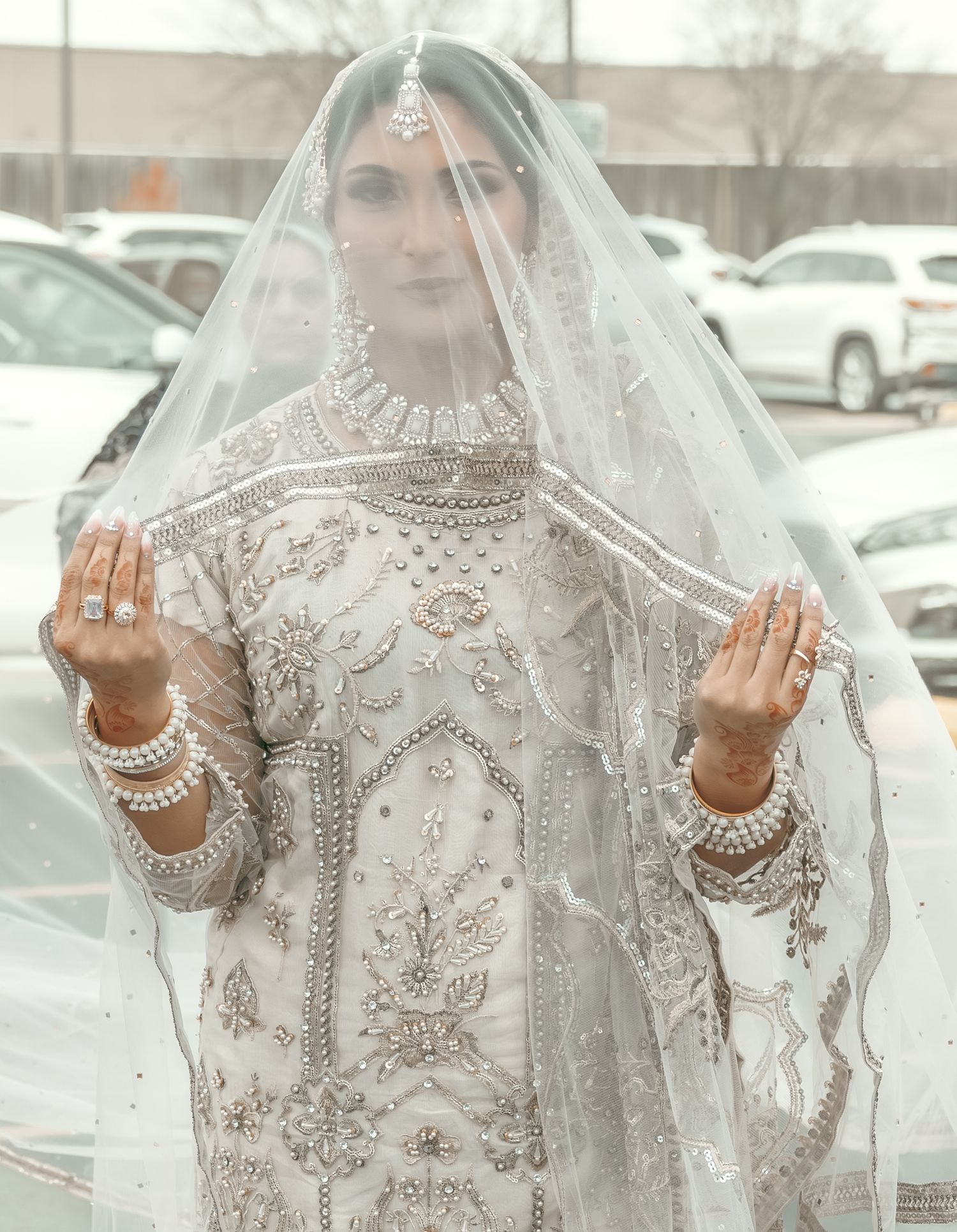 A South Asian bride in heavily embroidered white bridal wear holds her veil open in a parking lot.
