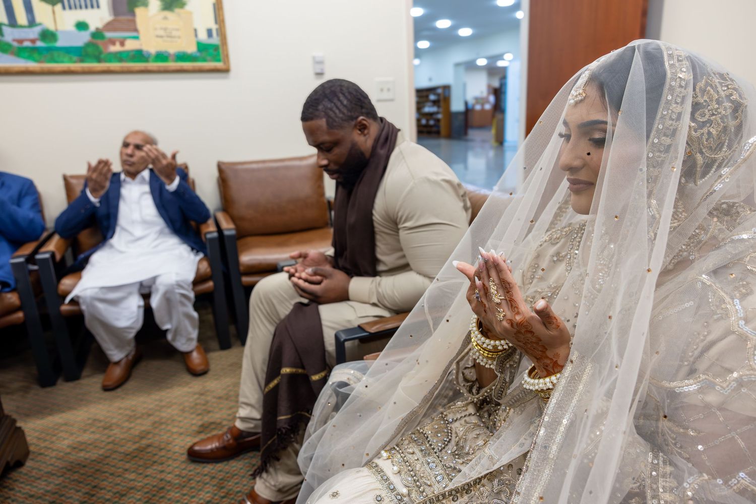 Bride in ornate white veil and groom in beige suit pray together during an Islamic Nikkah wedding ceremony.