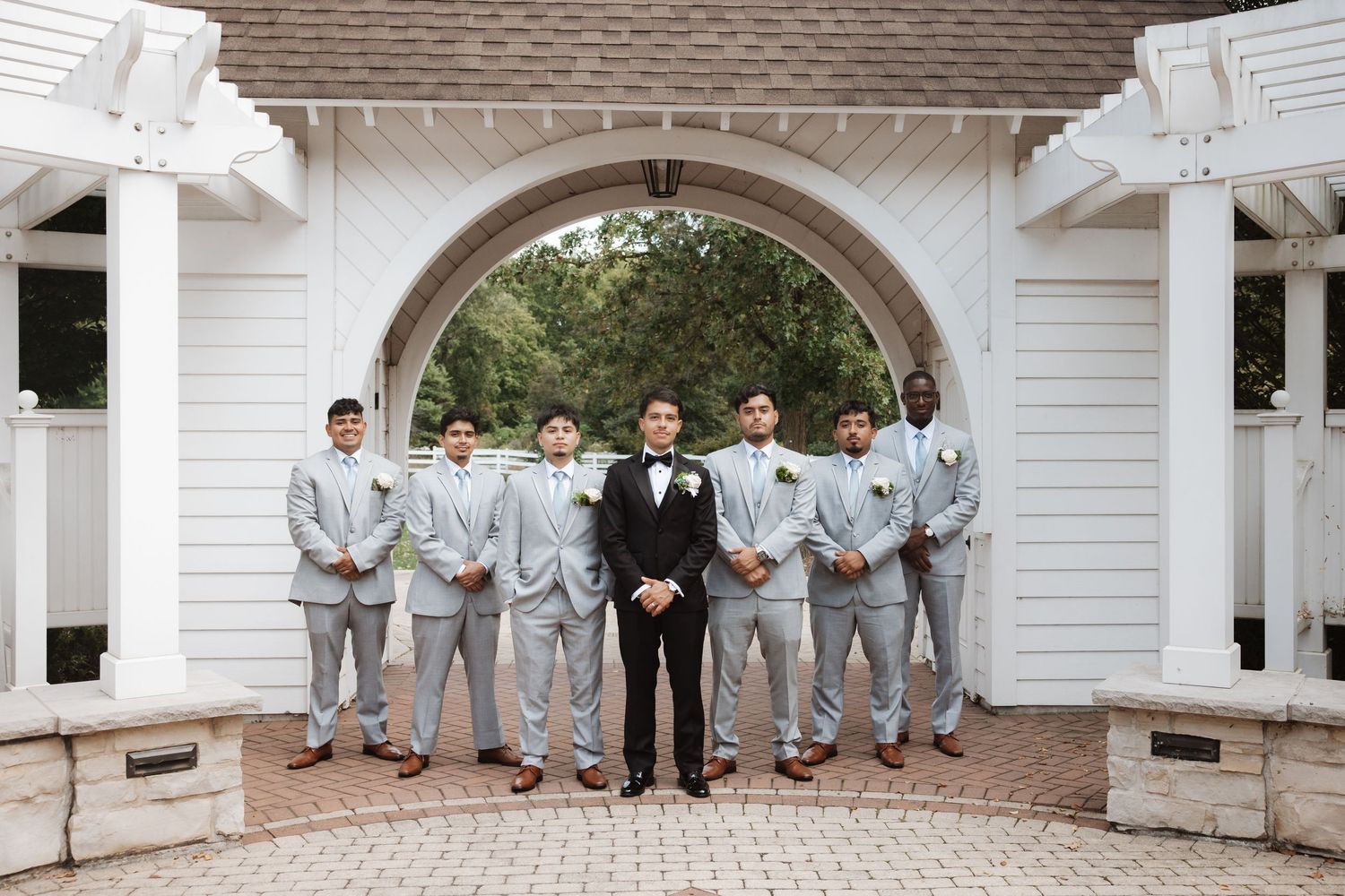 Groom in black tux stands centered with groomsmen in gray suits under arch.