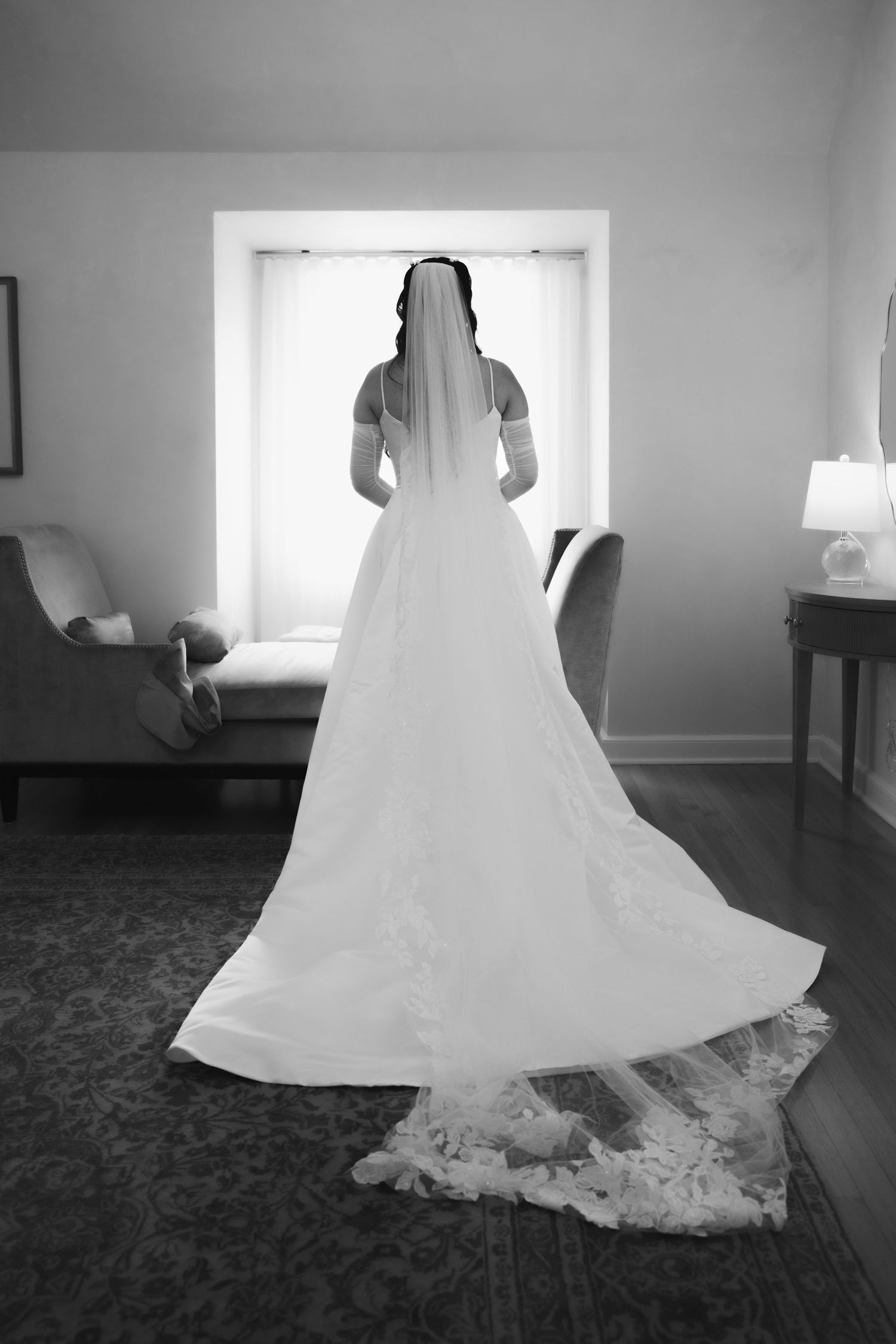 Elegant bride in long lace wedding gown and veil stands before window in black and white bridal portrait.