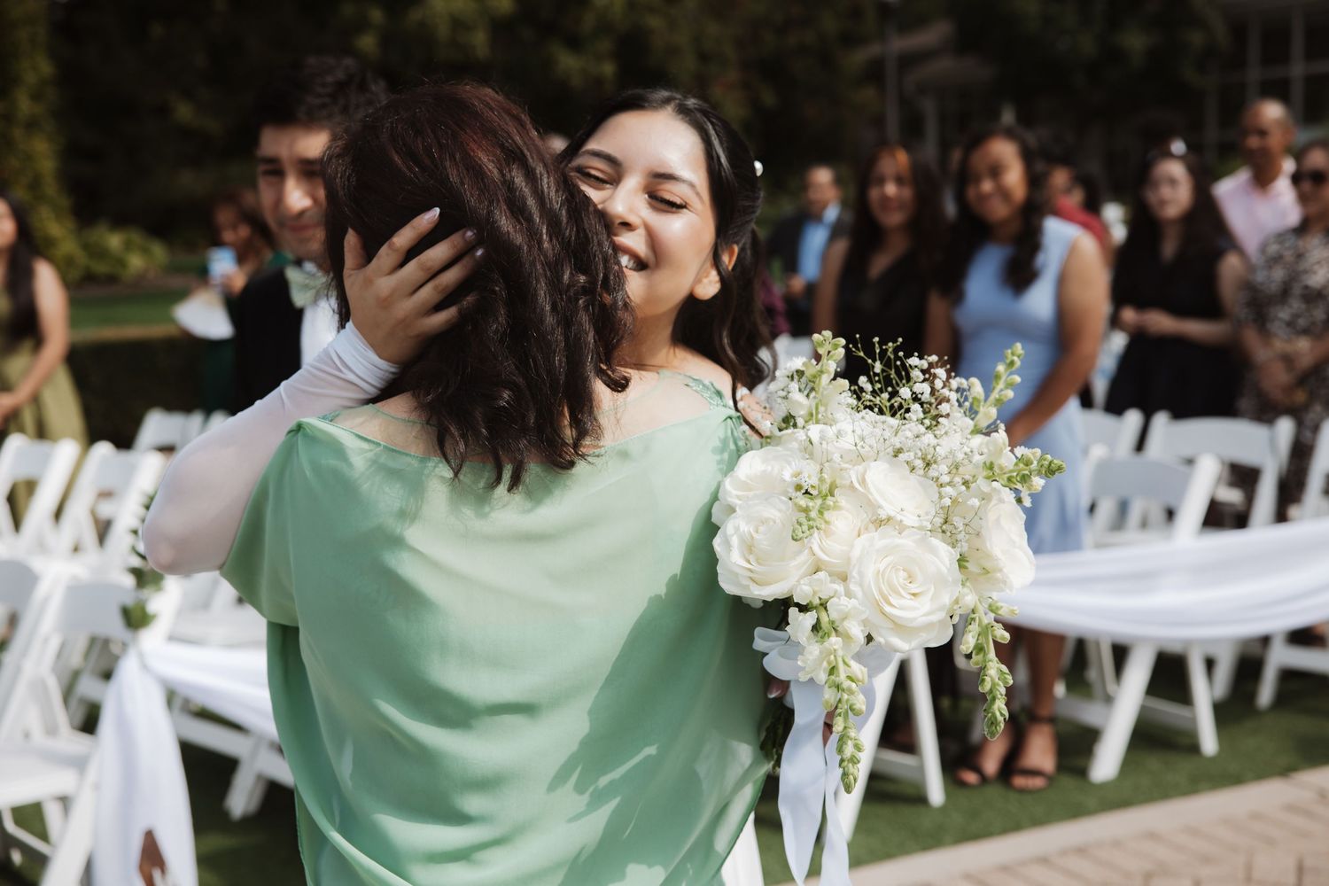 Bride embracing a guest in a mint dress while holding a white bouquet at an outdoor wedding.