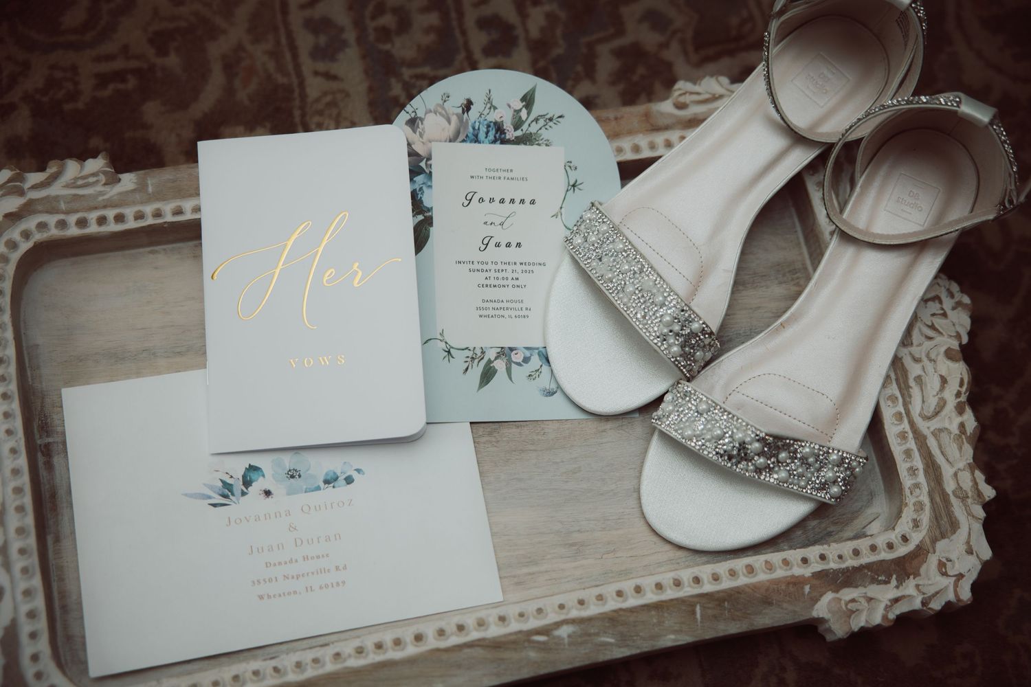 Wedding flat lay featuring glittery bridal heels, vow booklet, and floral invitation on ornate silver tray.