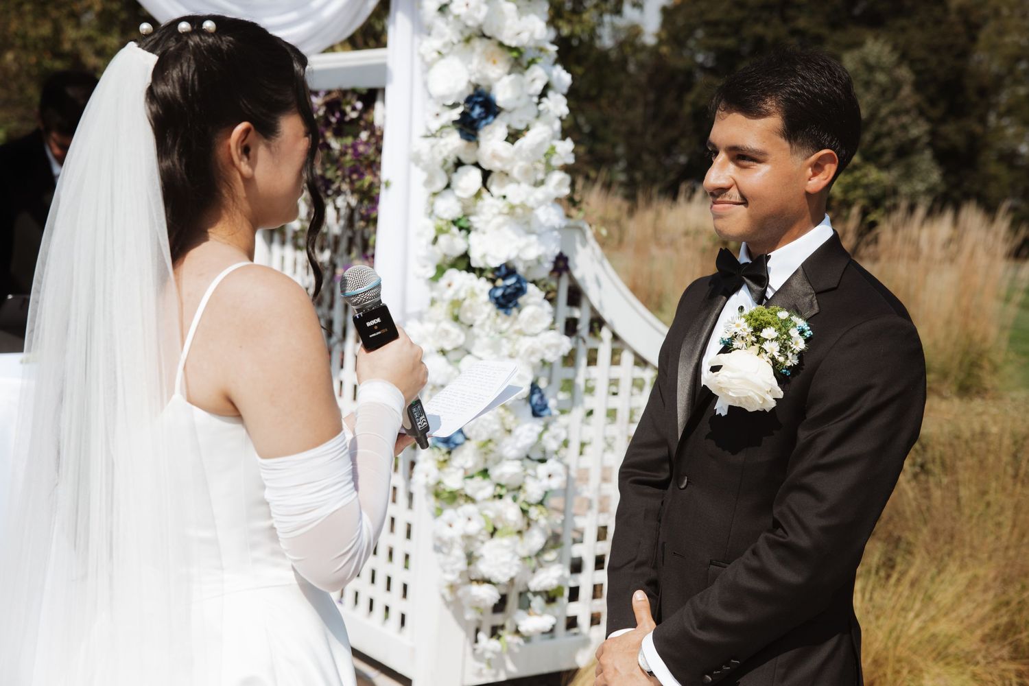 Bride in white veil reads vows to groom in black tuxedo at floral wedding arch outdoors.