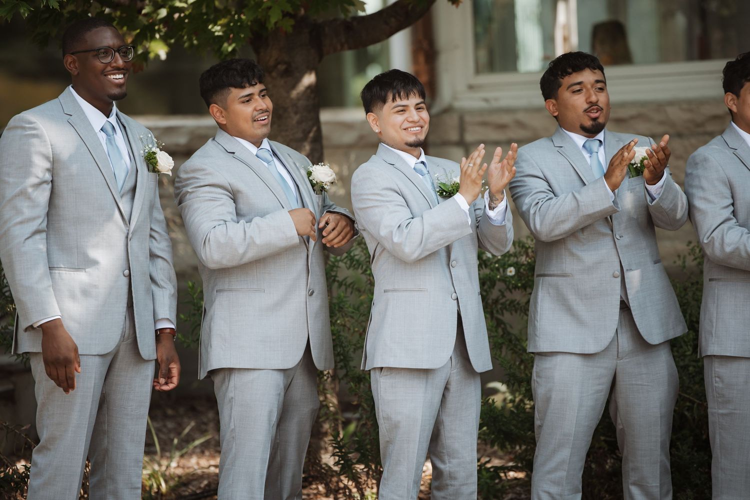 Four groomsmen in light gray suits stand together smiling and clapping during outdoor wedding celebration.