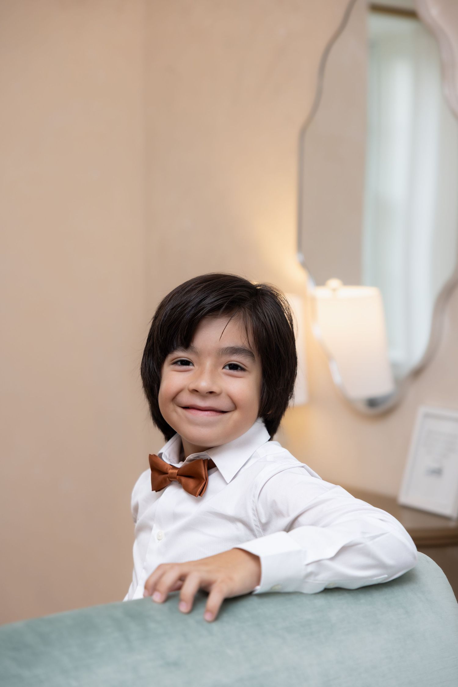 Smiling young boy in white shirt and brown bow tie posing at a formal wedding event.