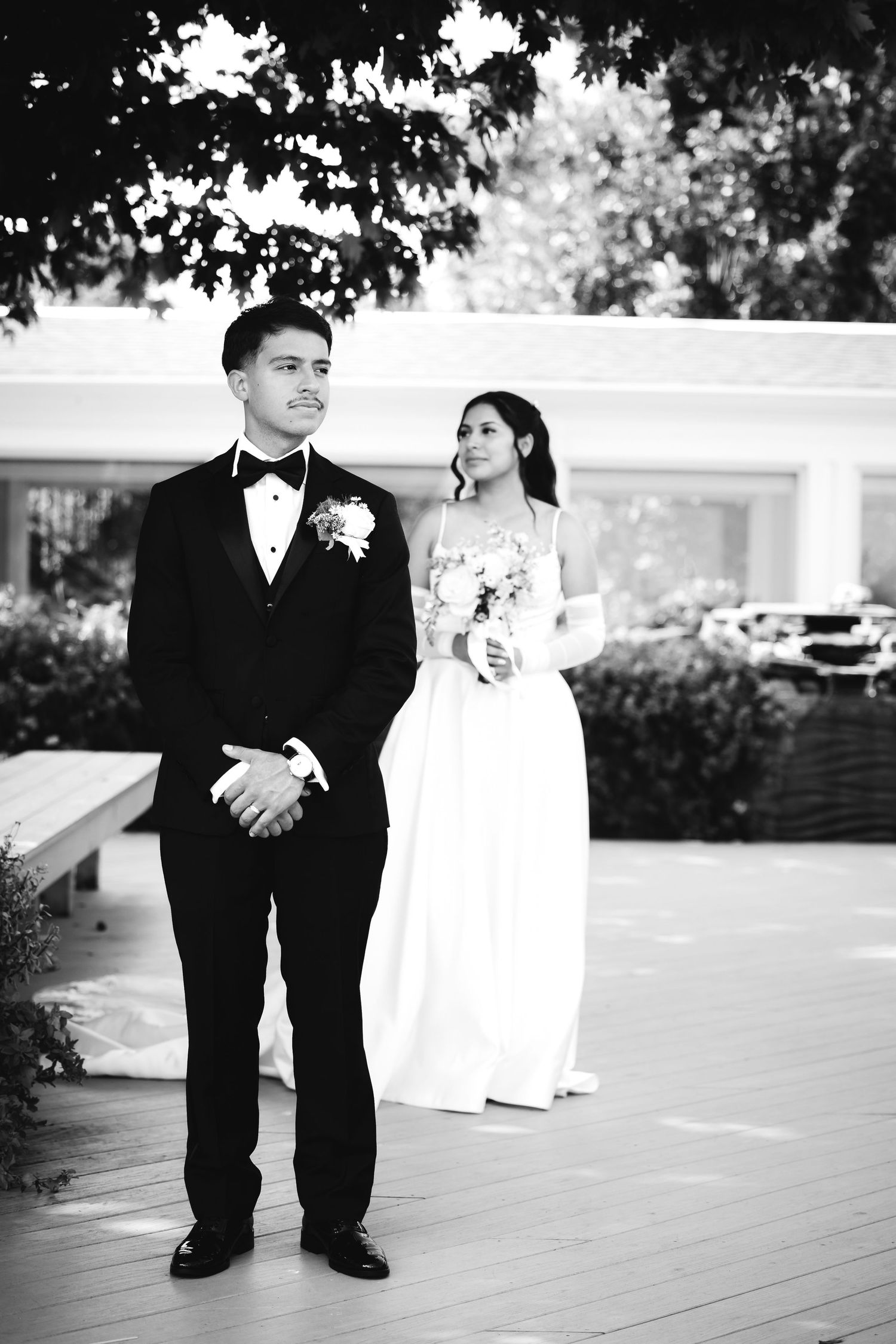 Black and white photo of groom in tuxedo standing before bride in white gown holding bouquet outdoors.