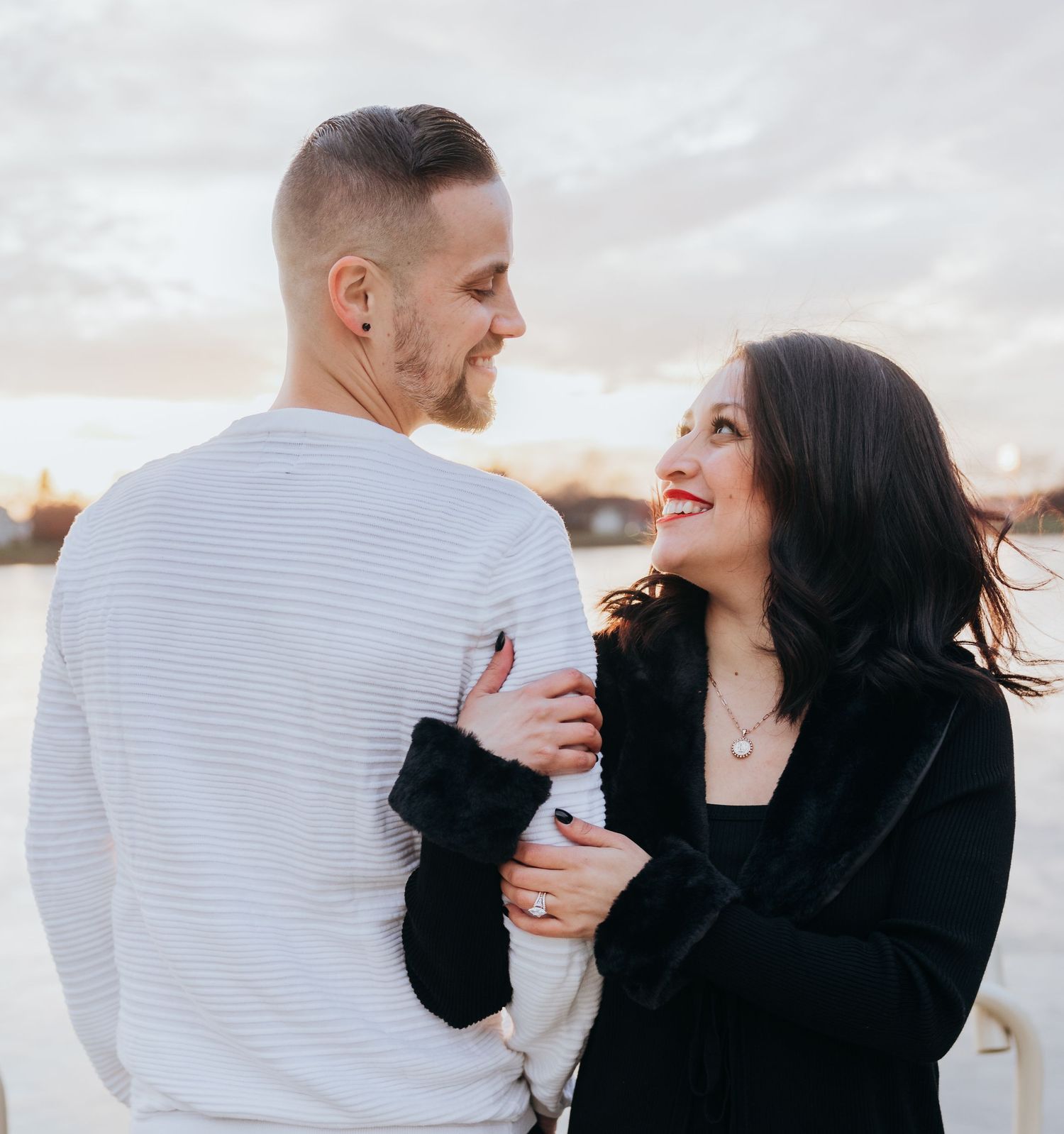 Romantic couple smiling at each other outdoors at sunset, woman in black jacket and man in white sweater.