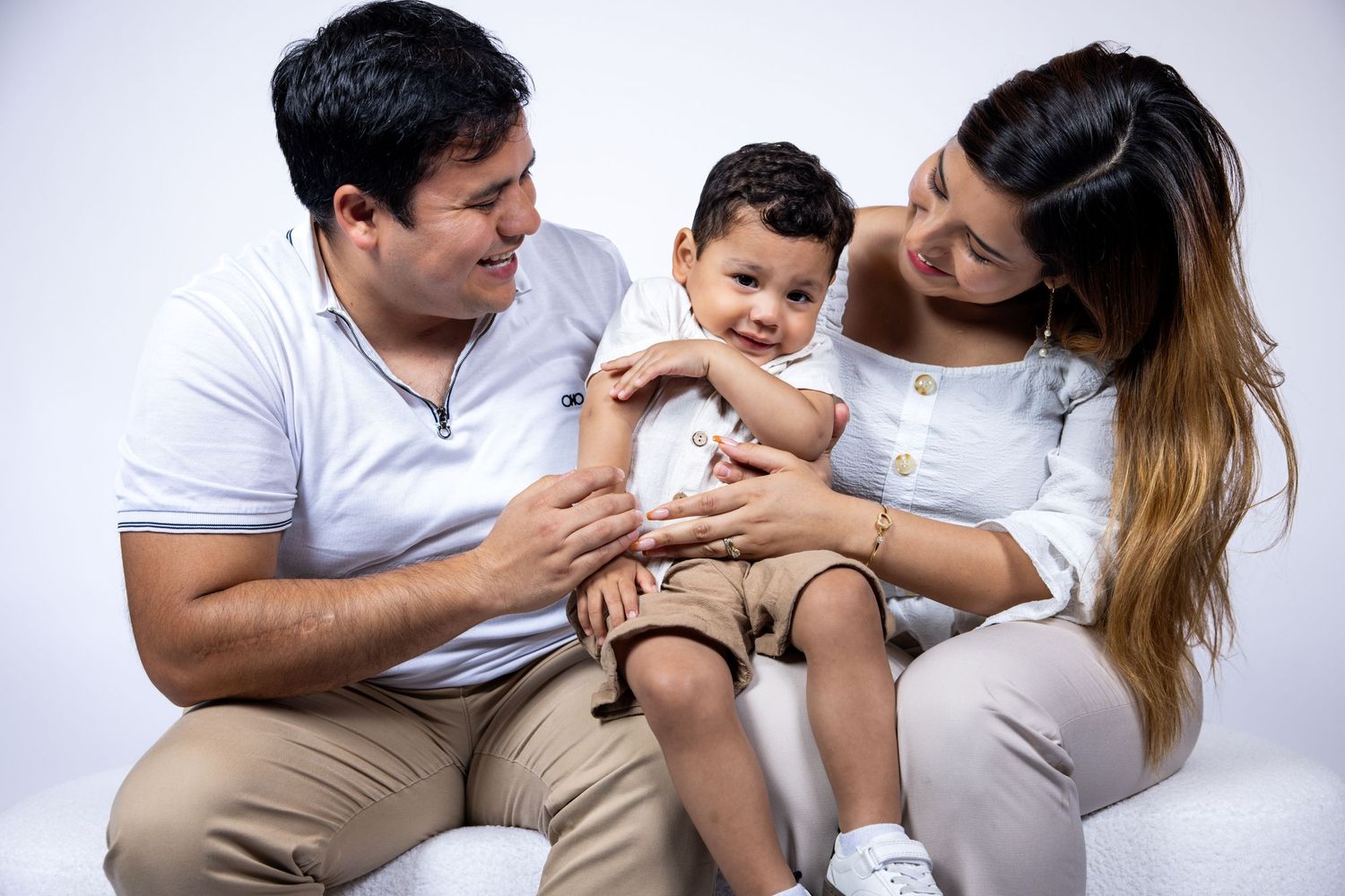 Happy family portrait with parents and baby boy in white and beige outfits on white background.