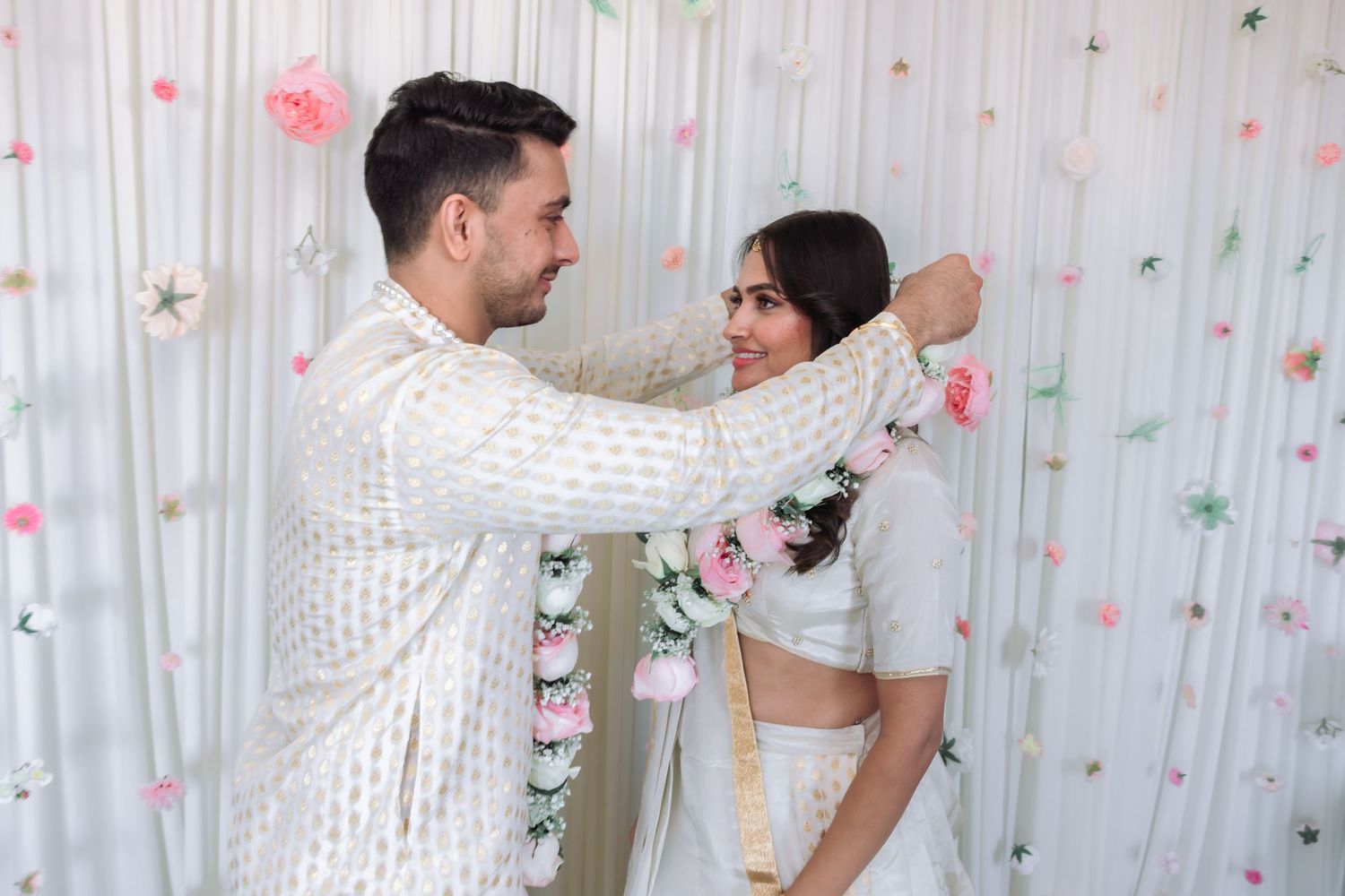 Indian groom adorns bride with pink floral garland during traditional engagement ceremony in white outfits.