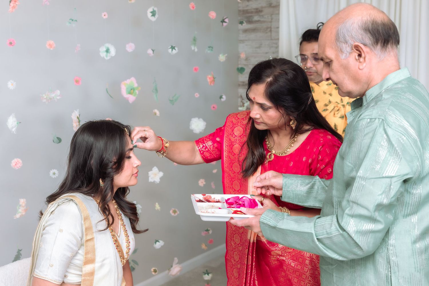 A woman in red saree applies tilak to a bride in white lehenga during an Indian wedding blessing ritual.