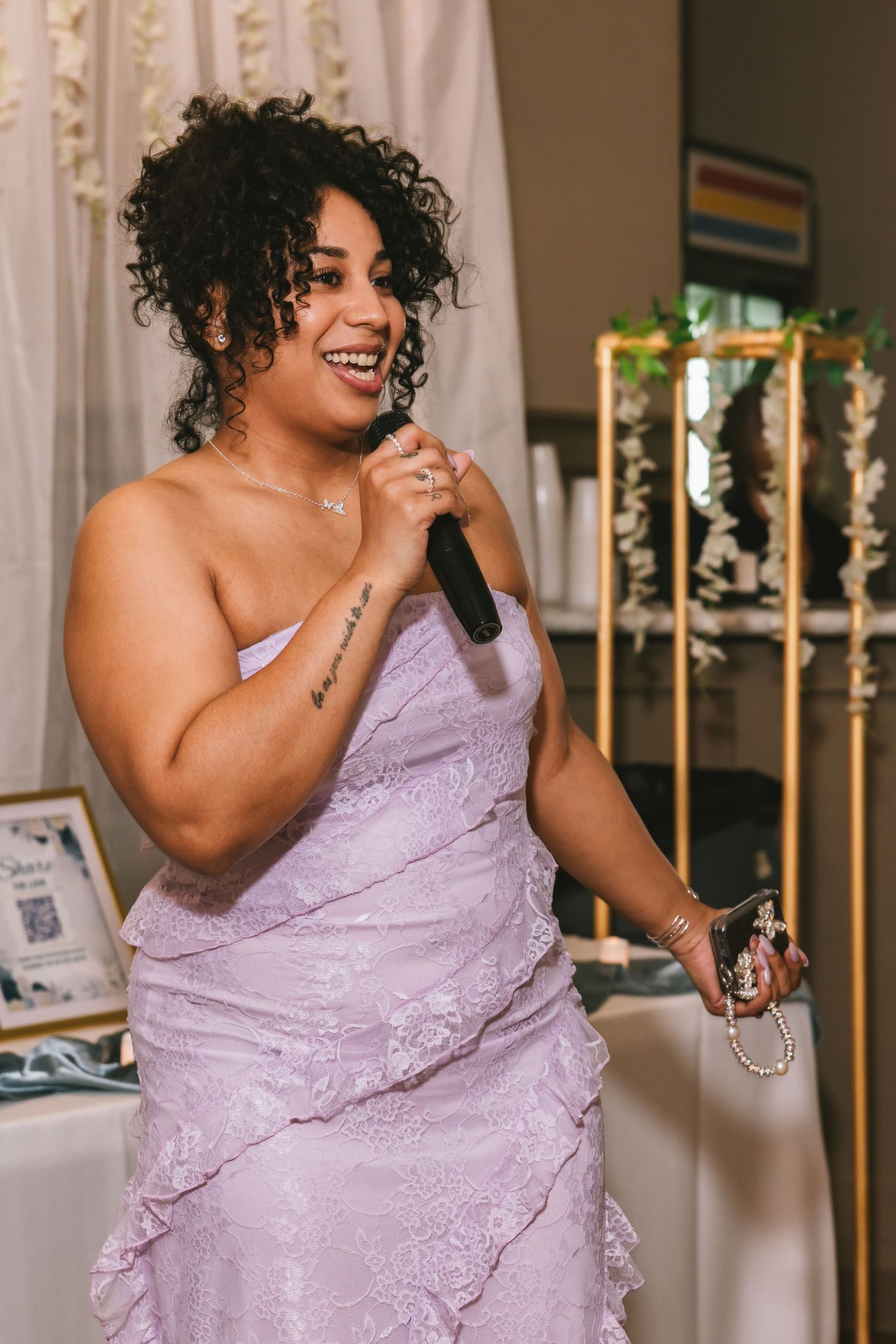 A woman in a lavender strapless lace dress holds a microphone while speaking at a wedding reception.