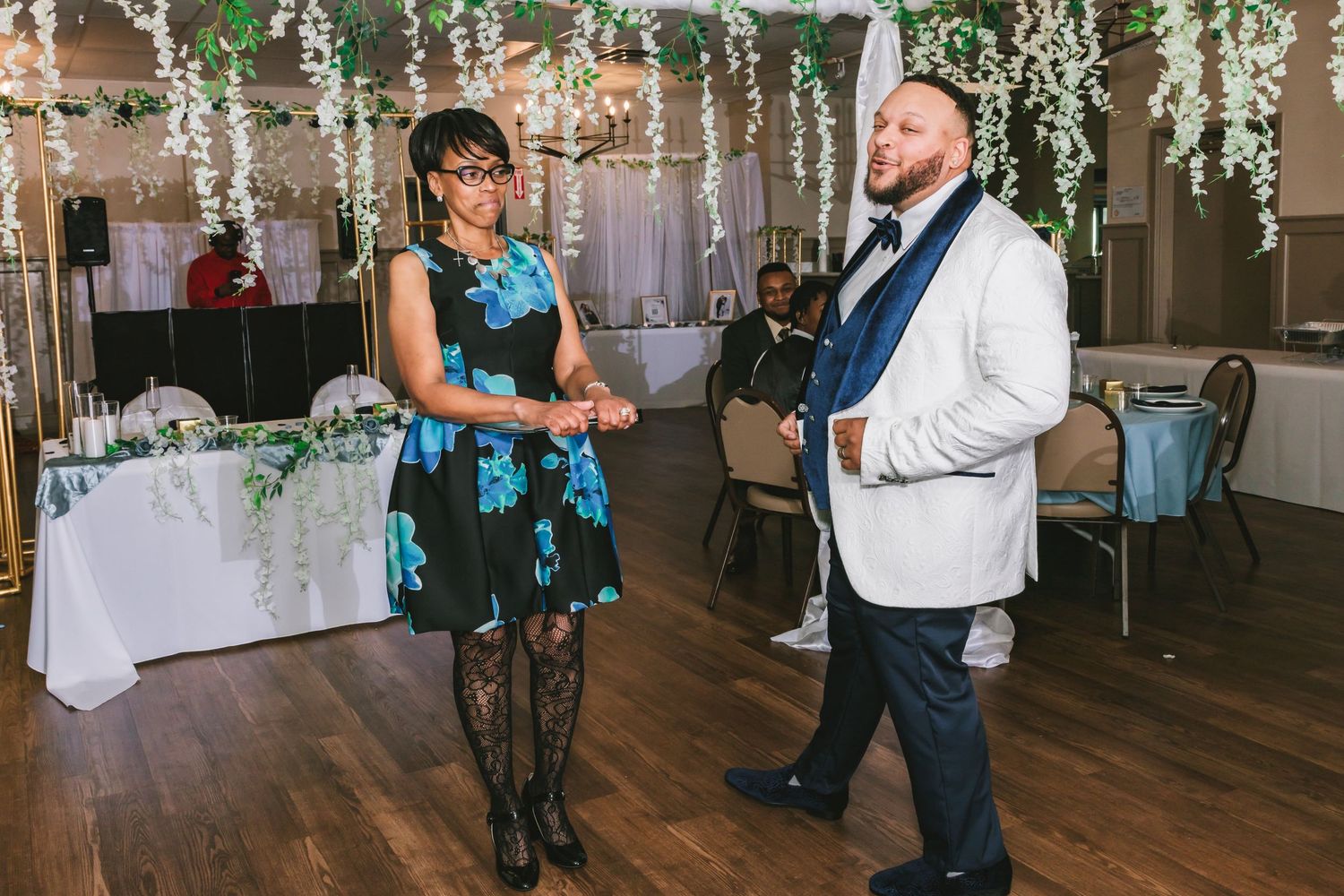 Groom in white jacket dances with a woman in floral dress at a festive wedding reception.